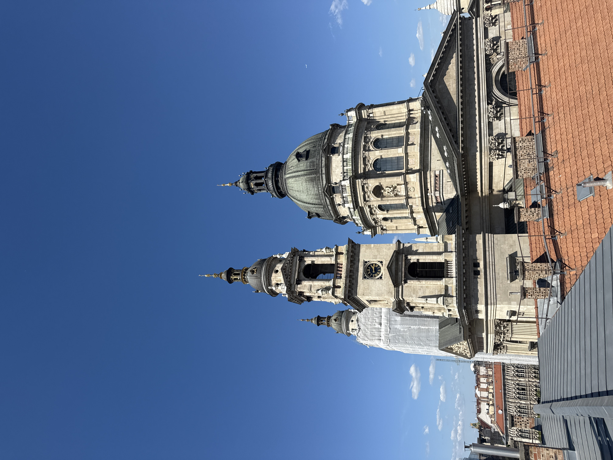 The image shows the Basilica of St. Stephen in Budapest, Hungary, from an elevated perspective. The building features prominent dome and towers, exhibiting detailed architectural elements. Part of the basilica is covered with scaffolding. The environment includes a clear blue sky with minimal clouds. The architecture in the foreground and background suggests an urban setting.