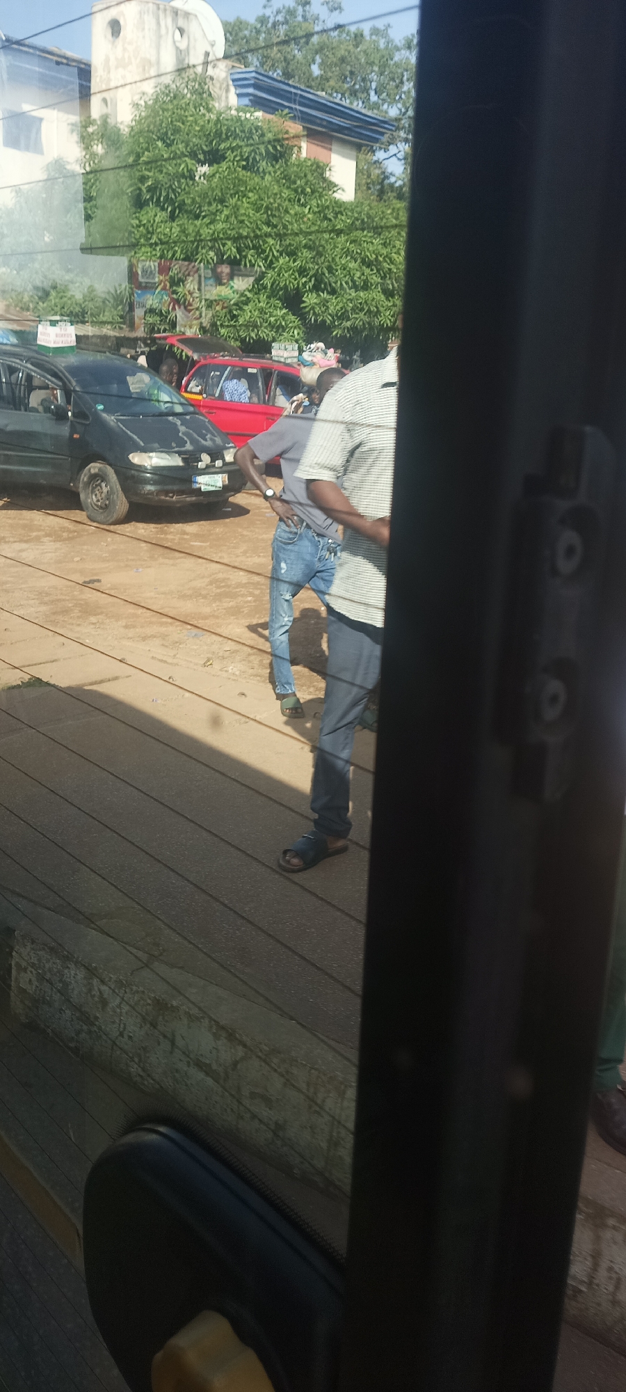 This outdoor photograph, taken in Jos, Nigeria, captures a vibrant street scene under bright daylight. The image is viewed through a window, evidenced by a dark frame on the right and bottom, and horizontal lines of reflection or dirt across the lower portion.
In the foreground, partially obscured by the window frame, are at least two men. One man, on the left, wears a grey t-shirt, ripped blue jeans, and green sandals, with a watch on his left wrist. He stands with his hands on his hips. Another man, closer to the right frame, wears a striped shirt and dark pants.
Behind the men, a black sedan (possibly a minivan like a Volkswagen Sharan) is parked or slowly moving. Its front bumper appears worn, and a light-colored license plate with dark characters is visible. Further back, a red car, possibly a taxi, is also visible. People can be seen inside both vehicles.
The background features lush green foliage, likely a large tree, possibly a mango tree, which partially obscures buildings. A white building with a blue trim or corrugated roof is visible behind the tree, topped with a satellite dish. A colorful poster with a person's face and some text is partially visible through the leaves of the tree, behind the black car.
The ground appears to be unpaved dirt or a worn road surface. The sunny conditions suggest midday or early afternoon, with clear skies. The overall scene depicts a typical bustling urban or semi-urban environment in Jos, with people and vehicles on the street.