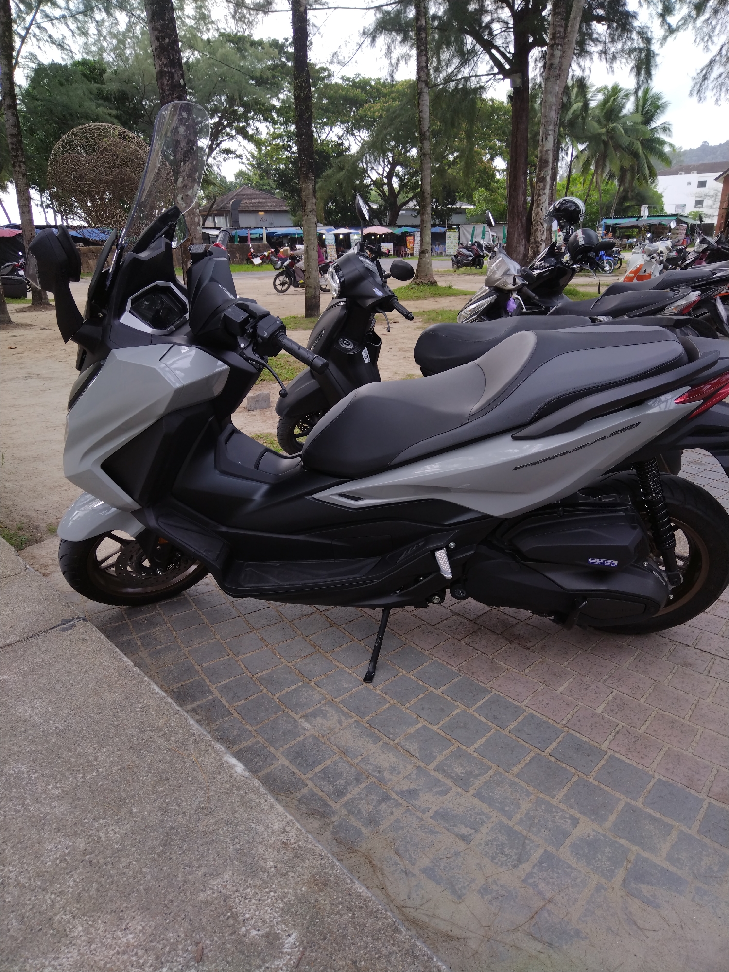 A light gray and black scooter is parked on a paved walkway next to a concrete step in Kamala, Thailand. The scooter is positioned at an angle, facing left, with its front wheel turned slightly. The scooter has a large windshield and a black seat. In the background, several other scooters are parked, along with trees and buildings. A large, circular, wire sculpture is visible among the trees on the left side of the image. The sky is overcast.