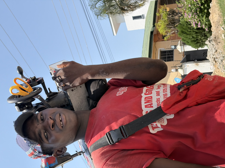 A young Black male stands in the foreground under a clear blue sky in Igboefon, Nigeria. He wears a red short-sleeved t-shirt with "B&B" and "TECH AND GADGETS" text, a distressed multicolored baseball cap, and two rings on his left hand. A dark-colored manual sewing machine, with prominent yellow scissors attached to its flywheel, rests on his right shoulder. A red shoulder bag with a black strap hangs across his chest. He looks directly at the camera with a neutral expression, revealing a small gap between his front teeth. The background features overhead utility wires. In the mid-ground, a light brown house with a green roof, security fencing, and a satellite dish is visible, adjacent to a more modern white building with a flat roof. Green foliage, including palm trees and flowering bushes, is present. The immediate foreground consists of rough ground with scattered debris.
