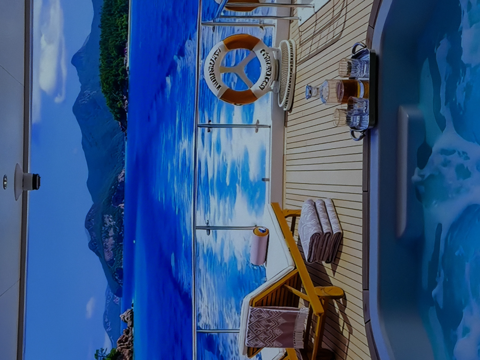 This image captures a vibrant digital display showcasing a luxurious deck scene, likely from a cruise ship or a high-end yacht, within an indoor setting. When viewed horizontally (the intended orientation of the displayed content), the scene unfolds with a picturesque landscape on the left and a detailed deck area on the right.
The left side of the display features a stunning natural vista: towering mountains covered in lush green foliage rise majestically from a serene, clear blue ocean, under a bright, cloudless sky. The water exhibits gentle ripples, enhancing the realistic depiction.
On the right, a meticulously rendered wooden deck is visible, featuring light-colored planks. A built-in jacuzzi or hot tub, filled with inviting bubbling water, occupies the far right. Adjacent to it, a black serving tray holds several glasses—some appearing to contain amber or clear liquids—and a decanter, suggesting an atmosphere of leisure and refreshments.
Further along the deck, a white and orange lifebuoy is mounted on a stand. The word "CRUISE" is clearly visible in black letters on its surface. Next to the lifebuoy stand, a small, white electric fan adds to the comfort of the virtual environment. A wooden lounge chair is positioned in the foreground, adorned with neatly folded white towels and a patterned textile. A pink and silver cylindrical object rests nearby. A sleek metal railing separates the deck from the simulated ocean.
The scene exudes a tranquil, daytime ambiance, suggesting perfect weather for relaxation. No people are visible within the digital scene. In the top left corner of the room where the photograph was taken, a white, wall-mounted device with a dark lens, likely a camera or sensor, is visible, indicating the real-world context of this impressive digital installation.