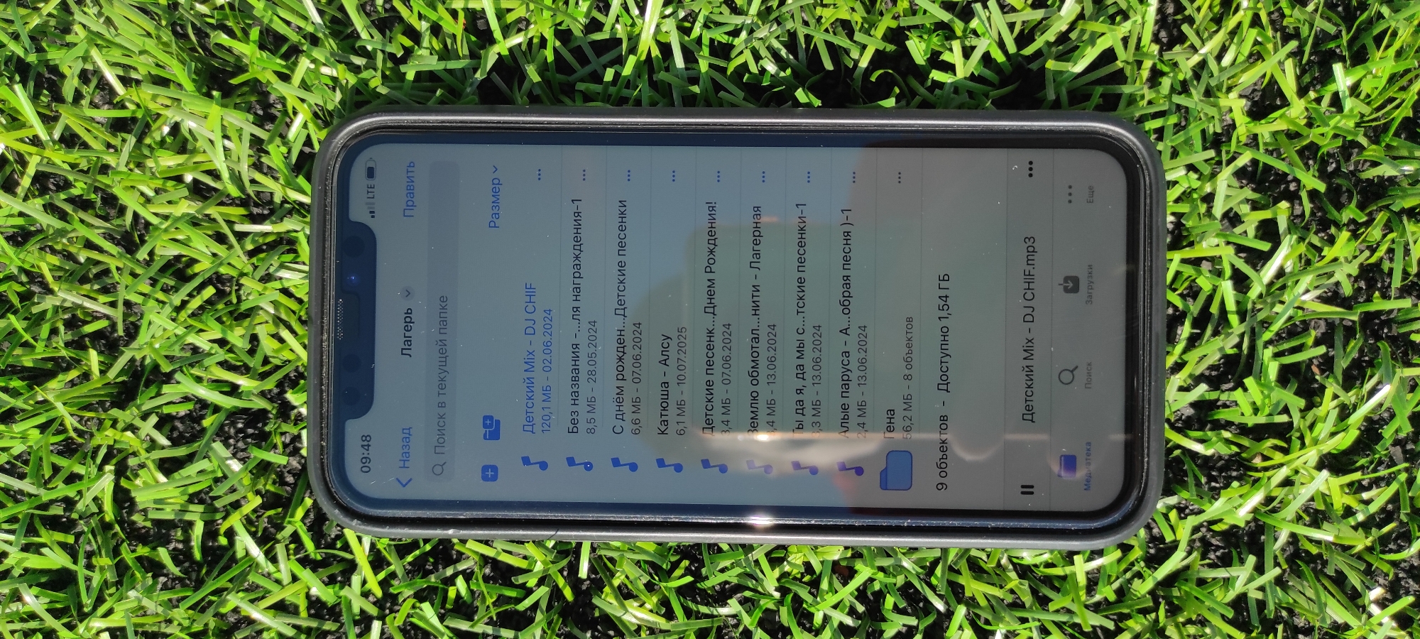 An iPhone is lying on artificial green turf, angled slightly upwards from the bottom left to the top right. The phone screen is on, displaying a file explorer or music application. The top bar of the phone shows "09:48" on the left and "LTE" and "ПРАВИТЬ" (Edit) on the right. Below this, "Лагерь" (Camp) is displayed, with a search bar labeled "Поиск в текущей папке" (Search in current folder). The list of files includes several entries with Russian names, dates, and file sizes. One entry is "Детский Mix - DJ CHIF" (Children's Mix - DJ CHIF) with a size of 120.1 MB and a date of 02.06.2024. Another entry is "Без названия" (Untitled) with a size of 8.5 MB and a date of 28.05.2024. Further down, there are entries like "Детские песенки" (Children's songs) and "Детский Mix - DJ CHIF.mp3". The bottom navigation bar shows icons for "Медиатека" (Media Library), "Поиск" (Search), and "Загрузки" (Downloads). The environment is solely the artificial turf, with no other elements visible. The image is not in Noviy, Russia, as there are no contextual clues to indicate this location.