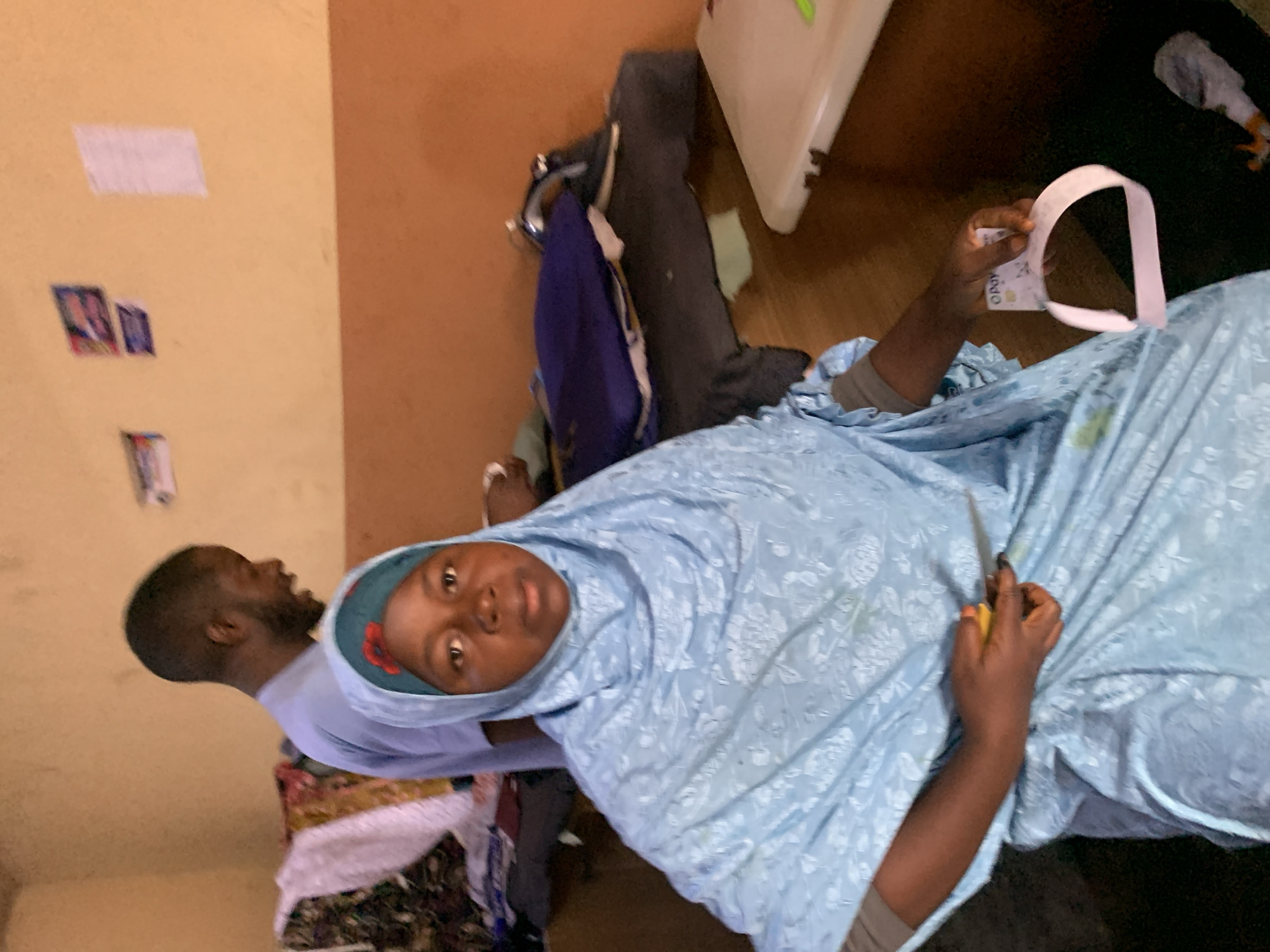 A woman wearing a light blue headscarf and gown is holding a pair of scissors in her right hand and a credit card in her left hand. To her left is a man wearing a light purple t-shirt. They appear to be indoors. The background includes a wall with papers affixed, various personal belongings, and what appears to be a white plastic object. The location is Jos, Nigeria.