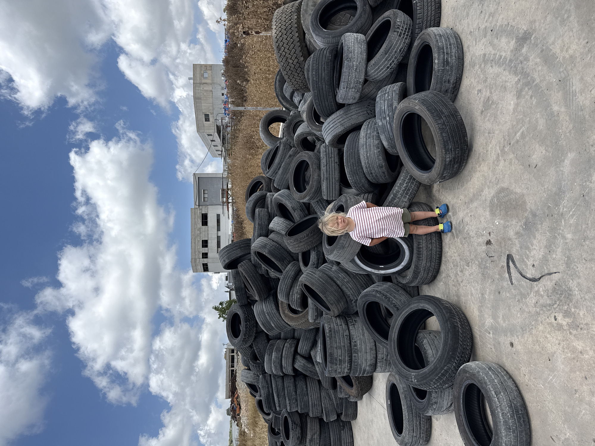 A young child stands in a scrapyard filled with stacked tires, located in Miilya, Israel. The child is wearing a striped t-shirt and shorts, and blue shoes. The tires are of various sizes and are piled high, creating a textured landscape. In the background, several industrial buildings are visible against a blue sky with scattered clouds. The ground is concrete, with some dark markings that appear to be oil stains or paint. The scene conveys a sense of industrial activity and the accumulation of discarded materials.
