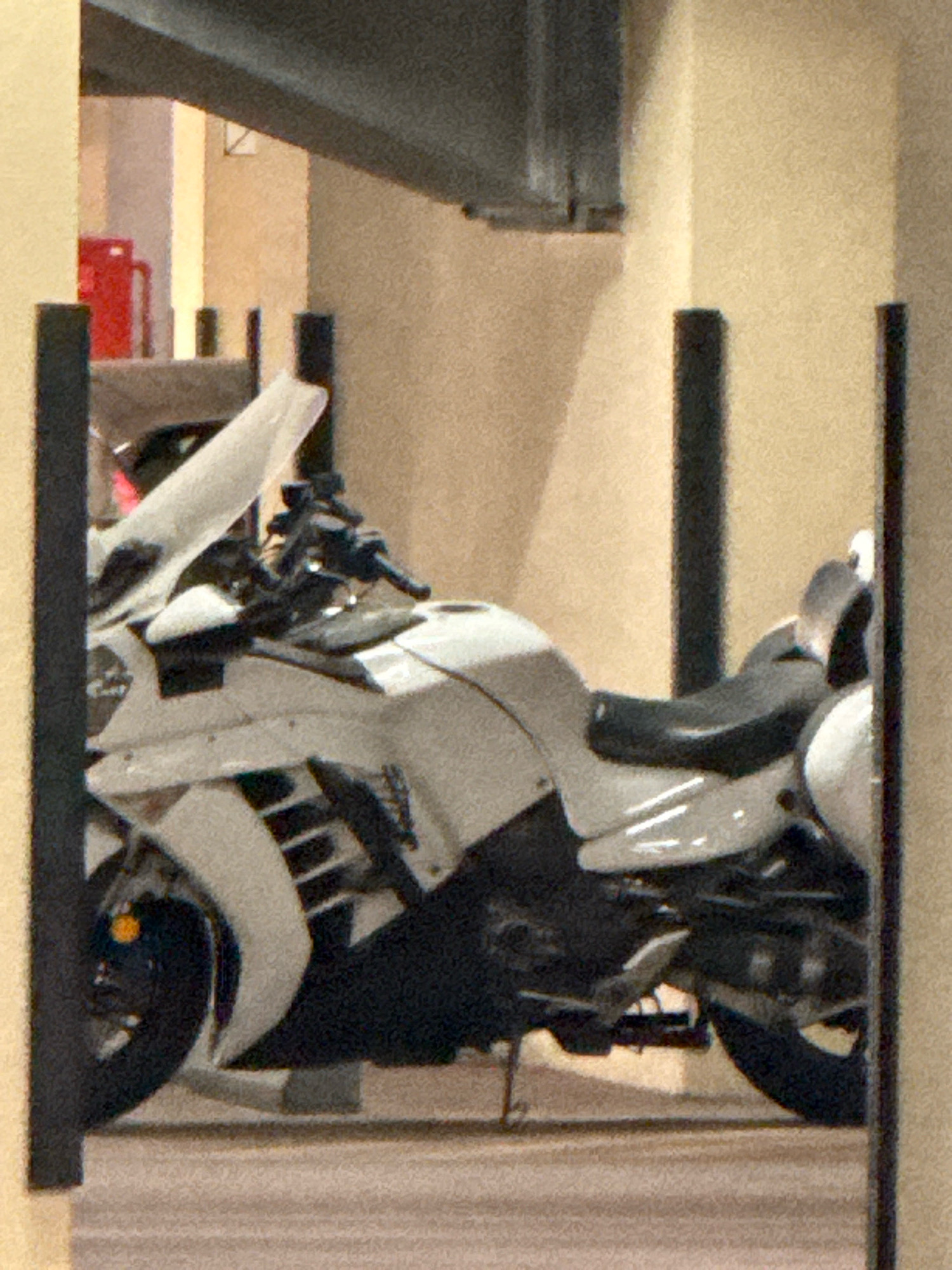The image shows a white motorcycle parked indoors, possibly in a parking garage or covered area. The motorcycle has a large windshield, a black seat, and chrome accents. The background consists of beige walls and a dark ceiling beam. There are vertical black poles in the foreground and midground, framing the motorcycle. In the background, a red fire extinguisher is visible. The image does not provide any specific indicators of its location in Dubai, United Arab Emirates.