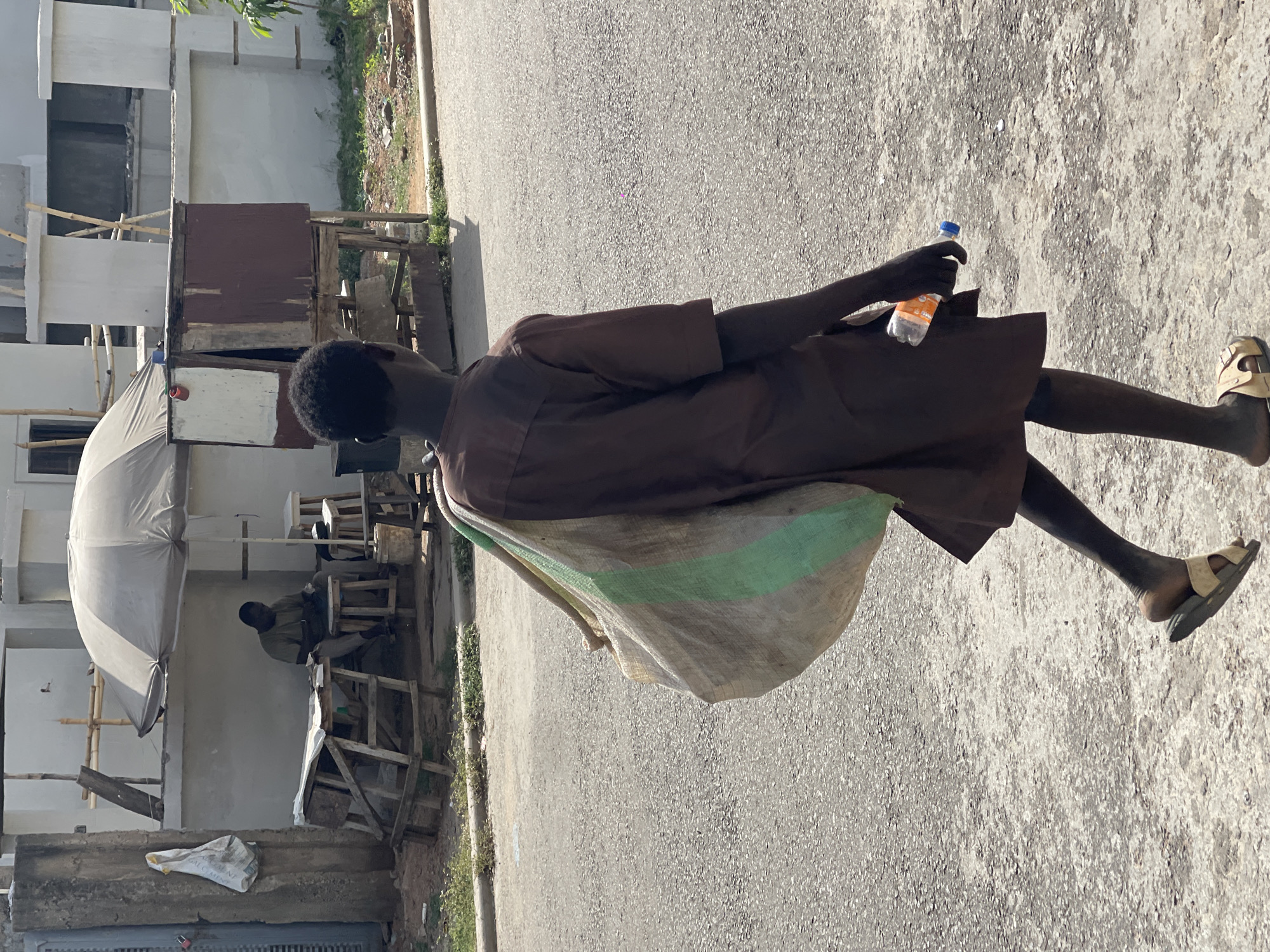 This image, taken in Gwarinpa, Nigeria, depicts a daytime outdoor scene on an asphalt road. The primary subject is a man, seen from behind and slightly to his right, walking towards the left of the frame. He is wearing a dark brown short-sleeved tunic and what appear to be dark trousers or bare legs, with light-colored, possibly beige, sandals on his feet. He has short, dark hair. Over his left shoulder, he carries a large, light-colored sack with a prominent green stripe running horizontally across its middle. In his right hand, he holds a transparent plastic bottle with an orange label and a blue cap.
In the background, to the left, are several informal structures. A large, light-colored umbrella or canopy provides shade over a seating area constructed from wooden planks and stools. Another man, wearing a lighter shirt, is visible sitting on one of these stools under the canopy. Further back, makeshift wooden stalls or shelters are visible, along with unpainted concrete buildings under construction, featuring bamboo scaffolding. A white bag, possibly for construction materials, is partially visible on the ground near these structures, with the word "CEMENT" faintly discernible. The road surface is granular asphalt, and a thin strip of grass and dirt borders it on the left. The scene is brightly lit by the sun, casting distinct shadows, indicative of a clear day.