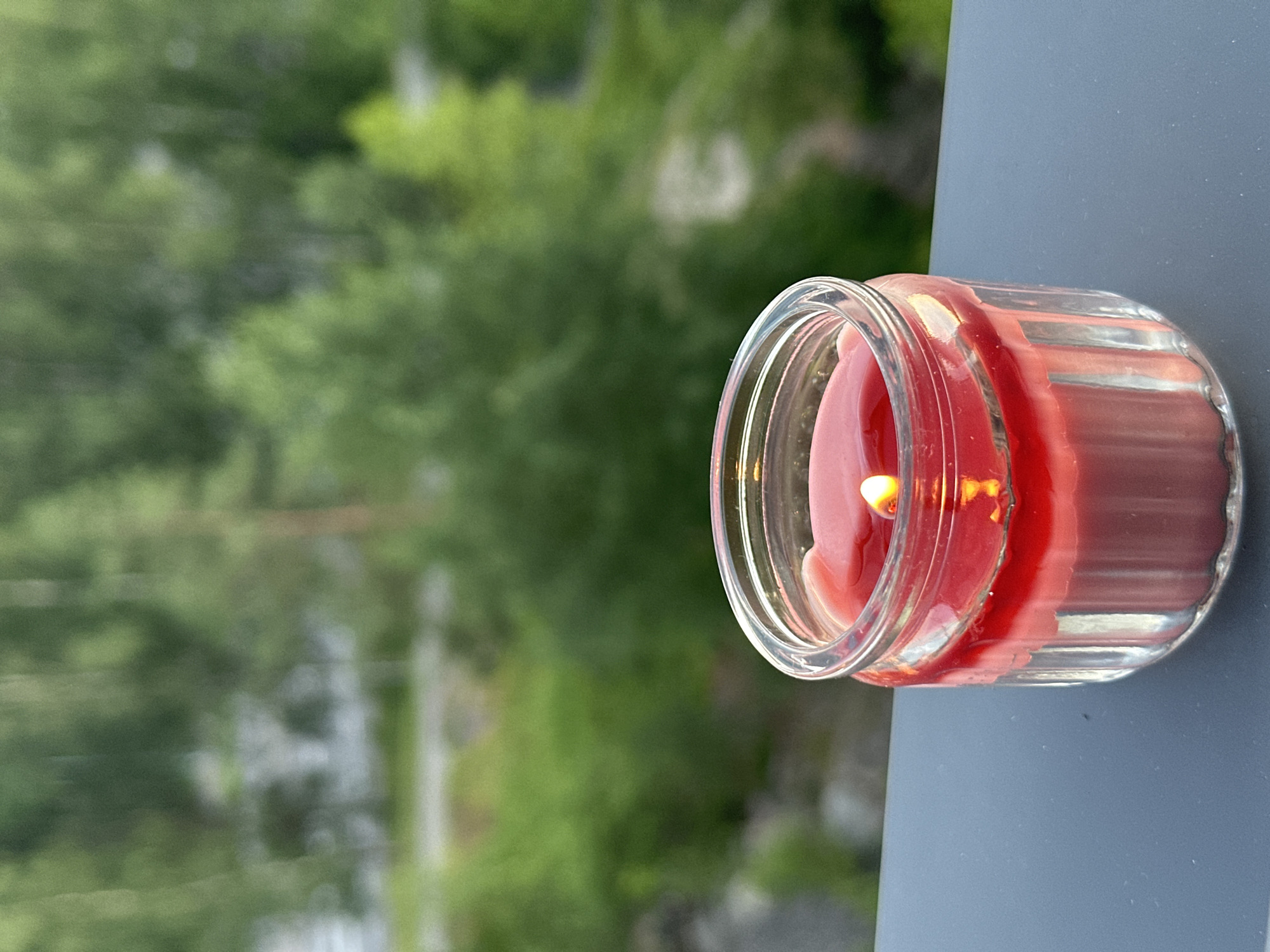 A lit red candle in a glass jar sits on a dark gray surface. The candle has a visible flame and the red wax is partially melted. The background is blurred and shows a natural scene with green trees and foliage, suggesting an outdoor location. The scene is not identifiable as being in a specific city.