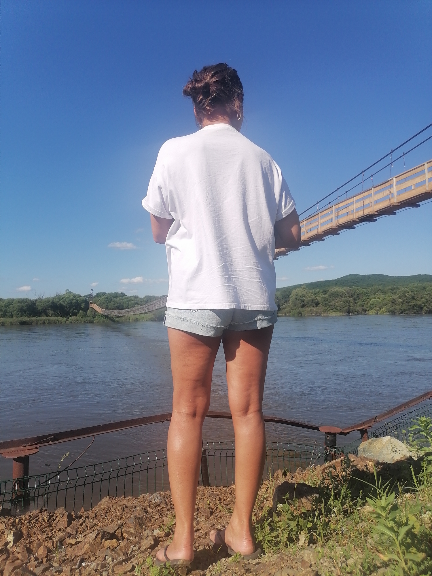 A woman stands with her back to the camera, facing a wide river under a clear blue sky. She has dark hair pulled up in a bun and wears a white t-shirt and light denim shorts. In the distance, two suspension bridges cross the river, surrounded by lush green trees and hills. The woman is standing on a rocky embankment with a metal fence and railing.