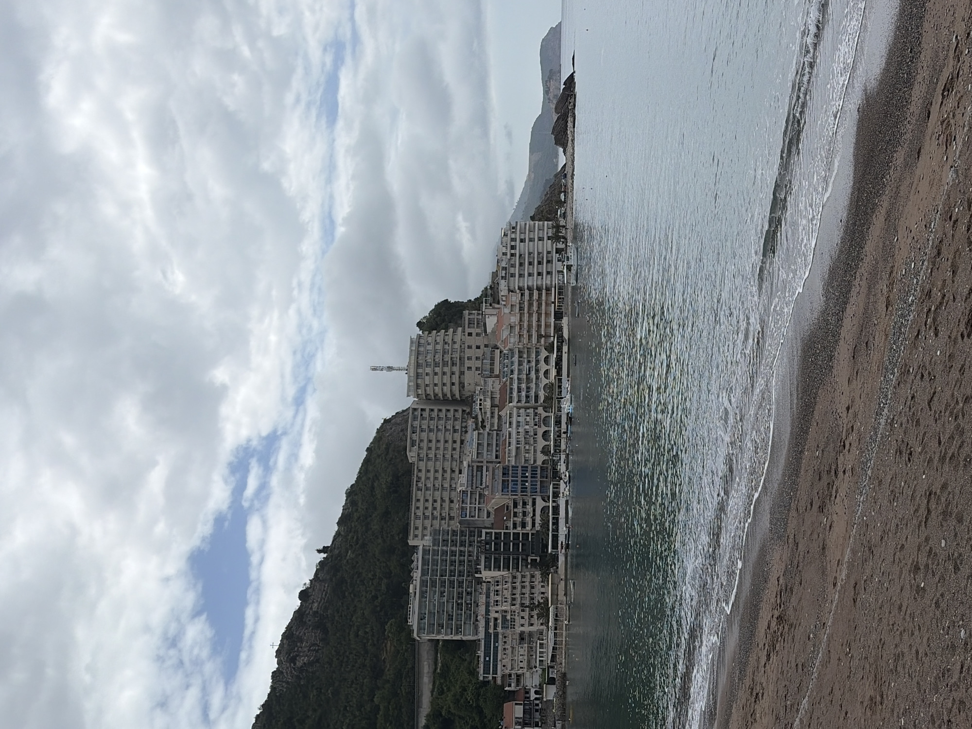 The image captures a coastal scene in Rafailovići, Montenegro. A series of modern, multi-story buildings are built on the side of a steep, green hill that descends to the shoreline. The buildings appear to be hotels or apartments. The sea stretches to the right of the frame, with gentle waves lapping onto a sandy beach. The sky is overcast with gray clouds, although some patches of blue are visible. The beach sand is dark and moist. The overall impression is of a densely built-up coastal resort area.