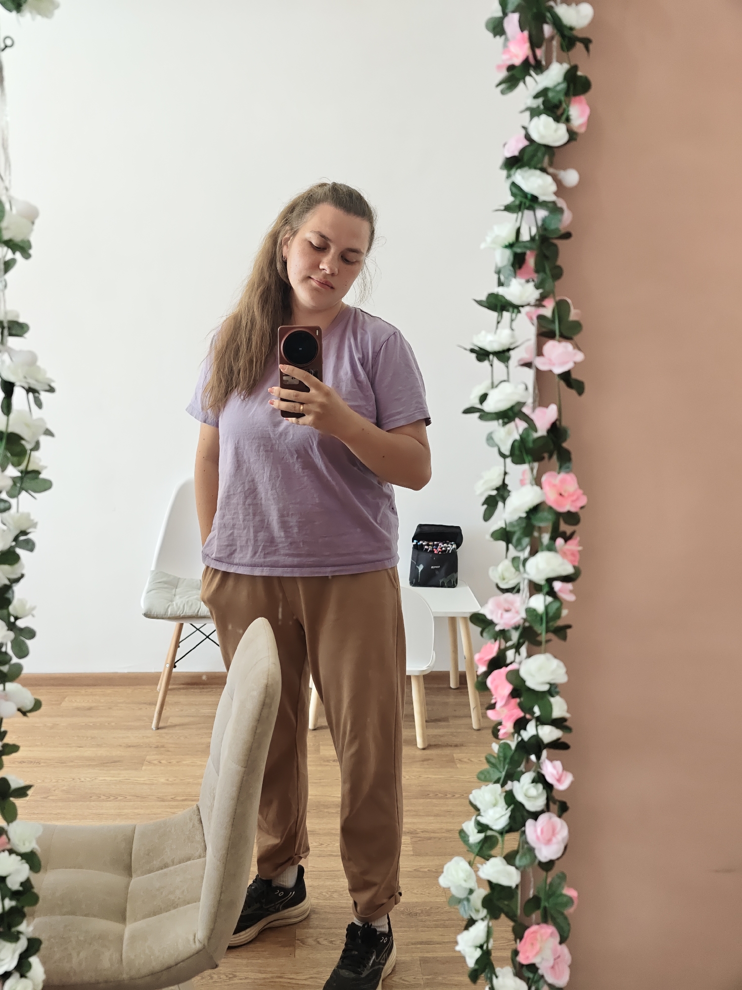 A woman is taking a selfie in front of a mirror decorated with white and pink flowers. The mirror reflects the room, which features a white wall, a light brown wall, a wooden floor, and chairs. One chair is white with wooden legs, and another is upholstered in beige. A black bag with what appears to be markers is on a white table. The woman is wearing a lilac t-shirt and tan pants, and black sneakers. Her hair is light brown. She is holding a reddish-brown smartphone. The setting appears to be indoors. No location or city can be identified from the image.