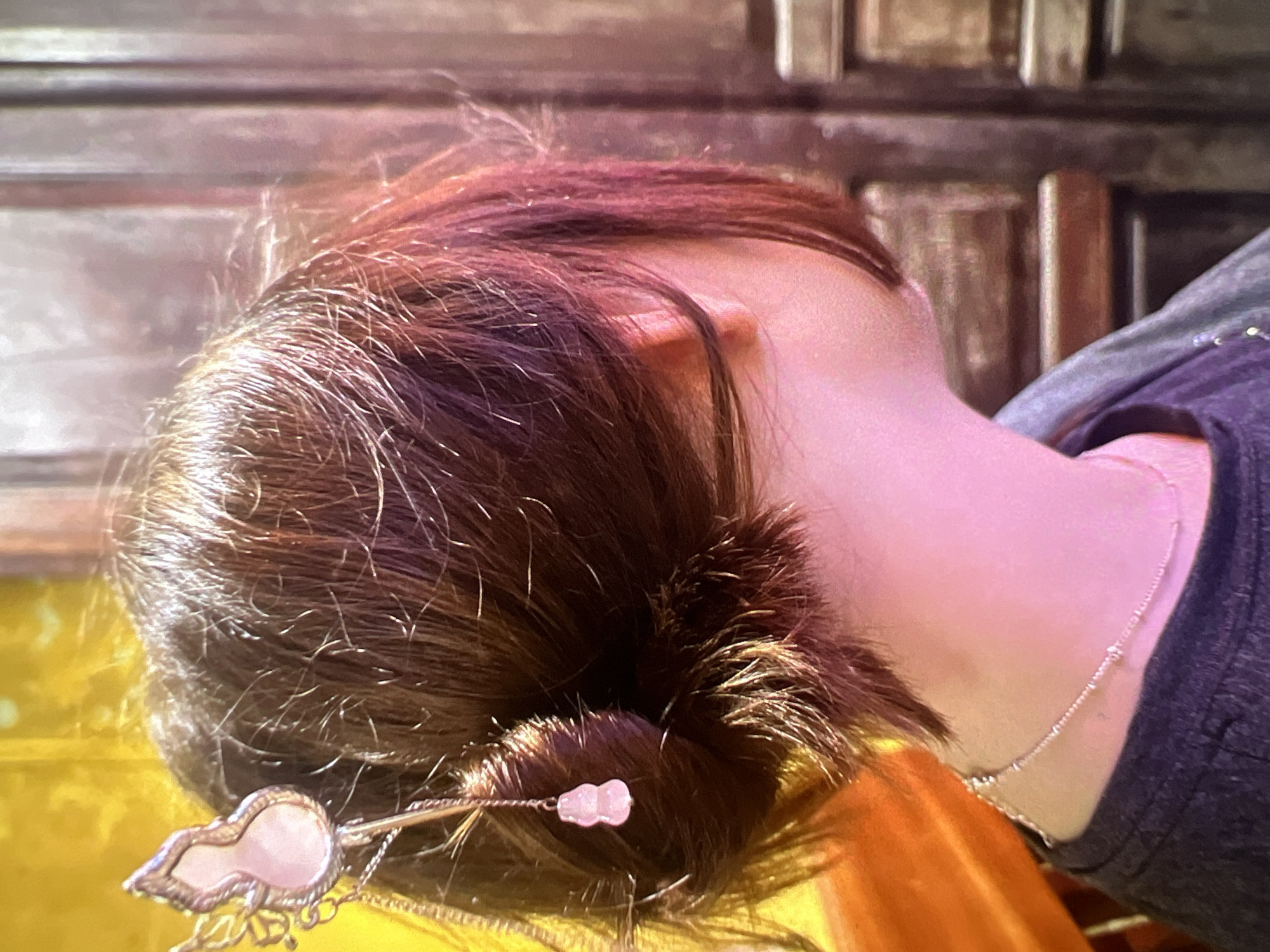 The image presents a close-up, top-down view of a person's head, neck, and shoulder, with the head tilted to the right. The person has dark brown hair, which is styled in a bun at the back of the head, showing some lighter reddish-brown strands and fine wisps. A decorative hair stick or hairpin is inserted into the bun. This accessory is silver-toned and features a pale pink or white cloud-like element, possibly made of mother-of-pearl or a gemstone, accompanied by small dangling chains and a tiny pink bead.
A thin, silver-toned necklace with small, evenly spaced beads or links is visible on the person's neck. The individual is wearing a dark grey or purple garment, likely a shirt, which covers the visible shoulder area.
The background is blurred, revealing warm-toned wooden panels or furniture. In the lower left portion of the image, a bright yellow or orange fabric or object is partially visible, which the person might be leaning against. The overall lighting is warm, casting a subtle pinkish hue on the skin and hair. No specific activity is discernible beyond the person appearing to be in a resting or leaning posture. There is no visible text in the image. The setting appears to be indoors.