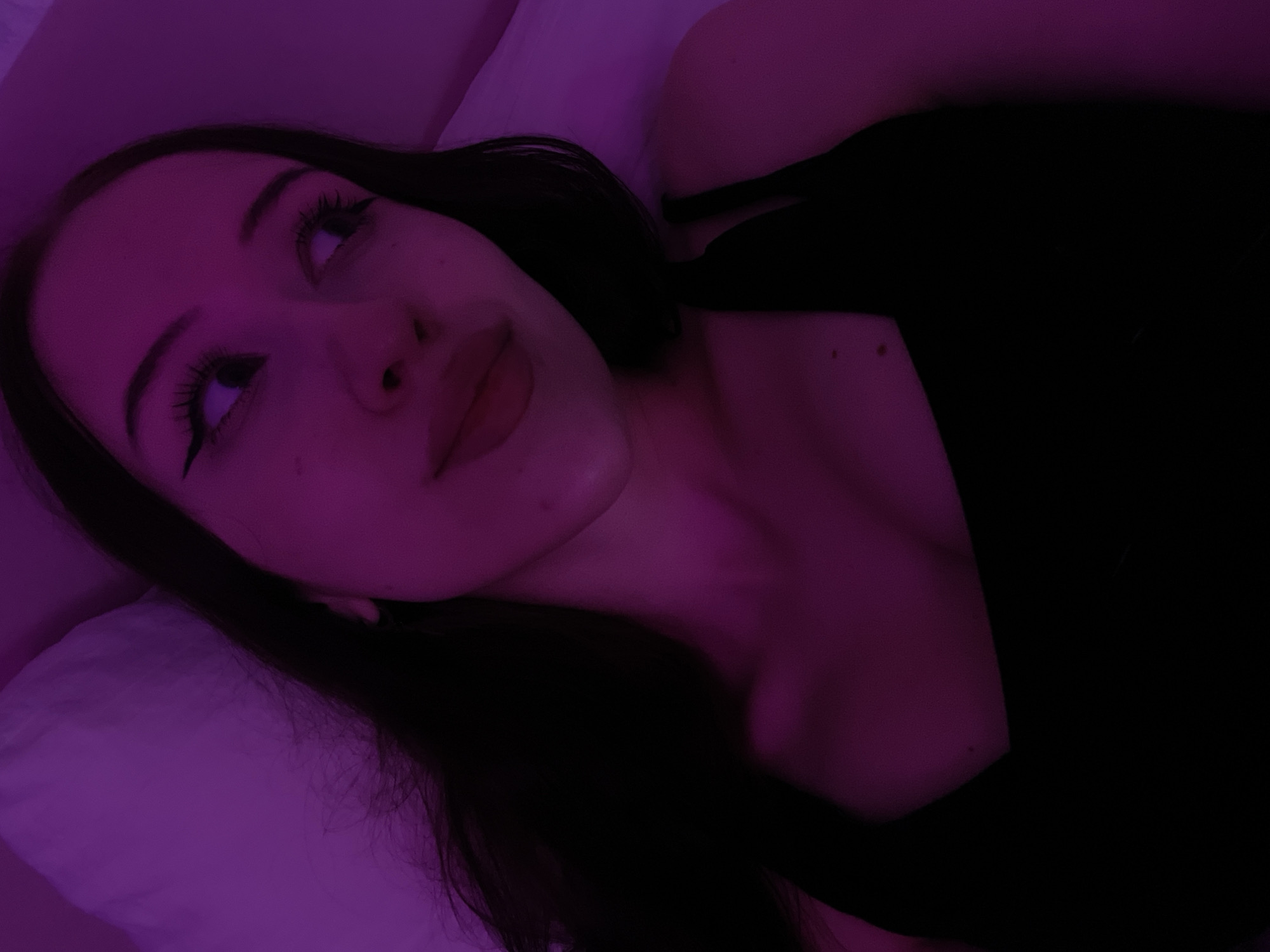 A young woman is lying on her side on a white pillow, illuminated by purple lighting. She has long dark hair, wears a black tank top, and is looking to her right. The image appears to be taken indoors, possibly in a bedroom. There is no indication of the specific location within Votkinsk, Russia.