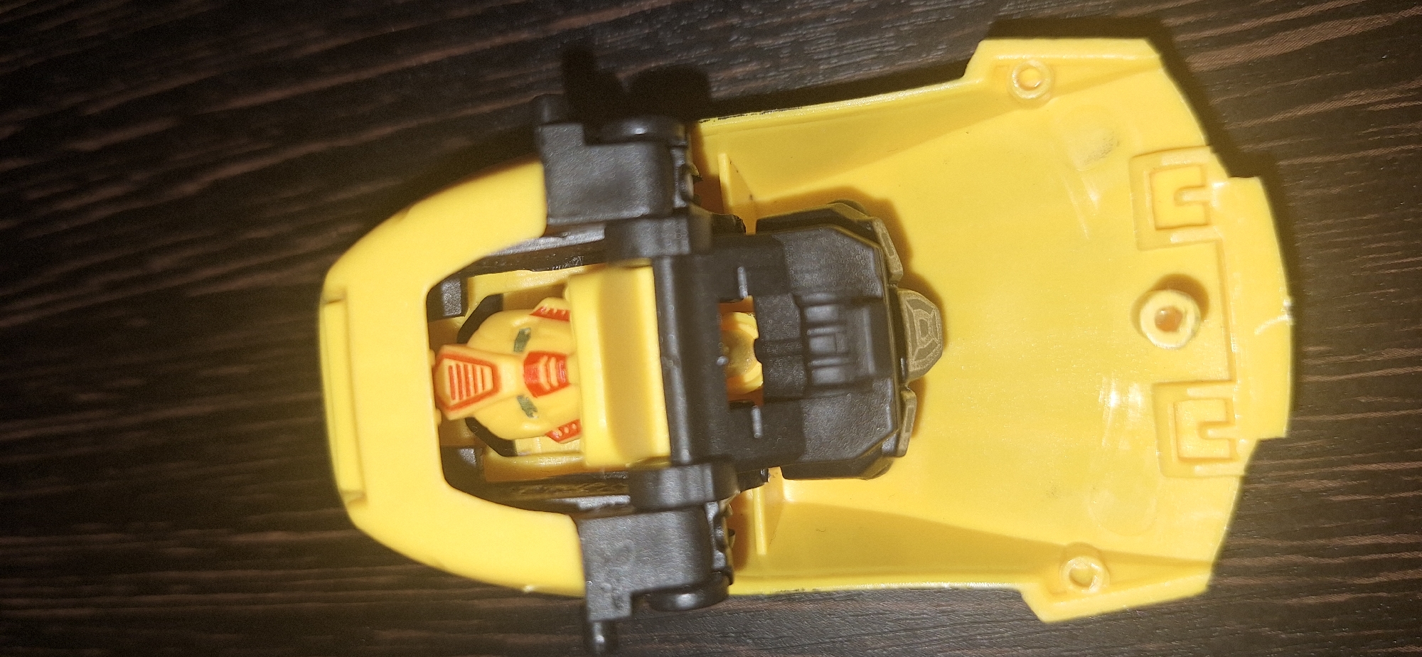 A yellow toy robot with red and orange accents is positioned in the center of the frame. The robot's head is visible within a yellow cockpit-like structure. The toy is placed on a dark brown surface with a visible wood grain pattern. The scene is captured from a top-down perspective. There is no indication of the location in Bila Tserkva, Ukraine.
