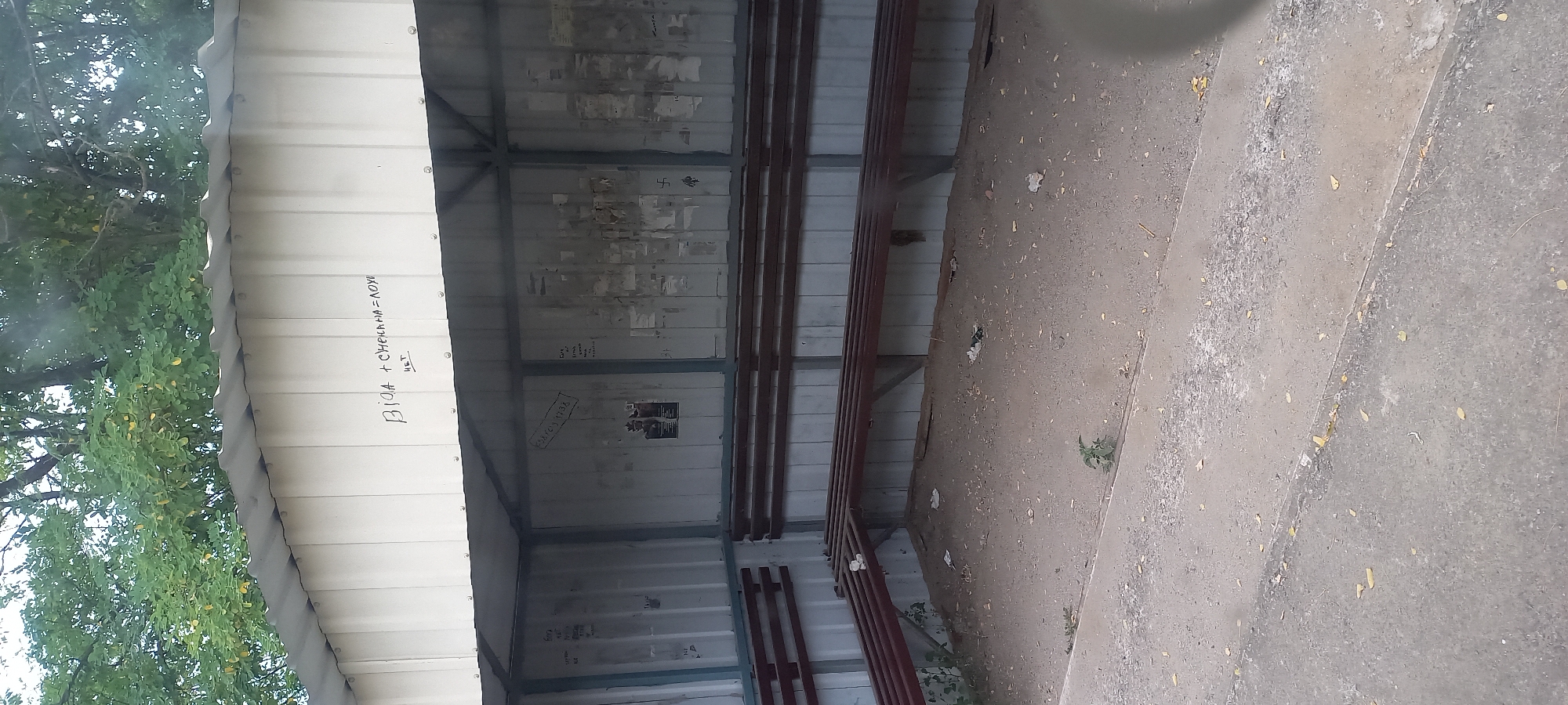 A bus stop shelter with a corrugated metal roof and siding is depicted. The shelter has two rows of slatted wooden benches. The ground inside the shelter is sandy and littered with debris. Graffiti is present on the exterior wall of the shelter, including the inscription "ВІД + СНЕЖАНА = ЛЮБЛЮ" (VIO + SNEZHANA = LOVE) and "НЕТ" (NO). There are also various posters and markings on the interior walls, some of which are illegible. The bus stop is located outdoors, with trees visible in the background. The image appears to be taken from a slightly elevated position, looking towards the bus stop entrance.