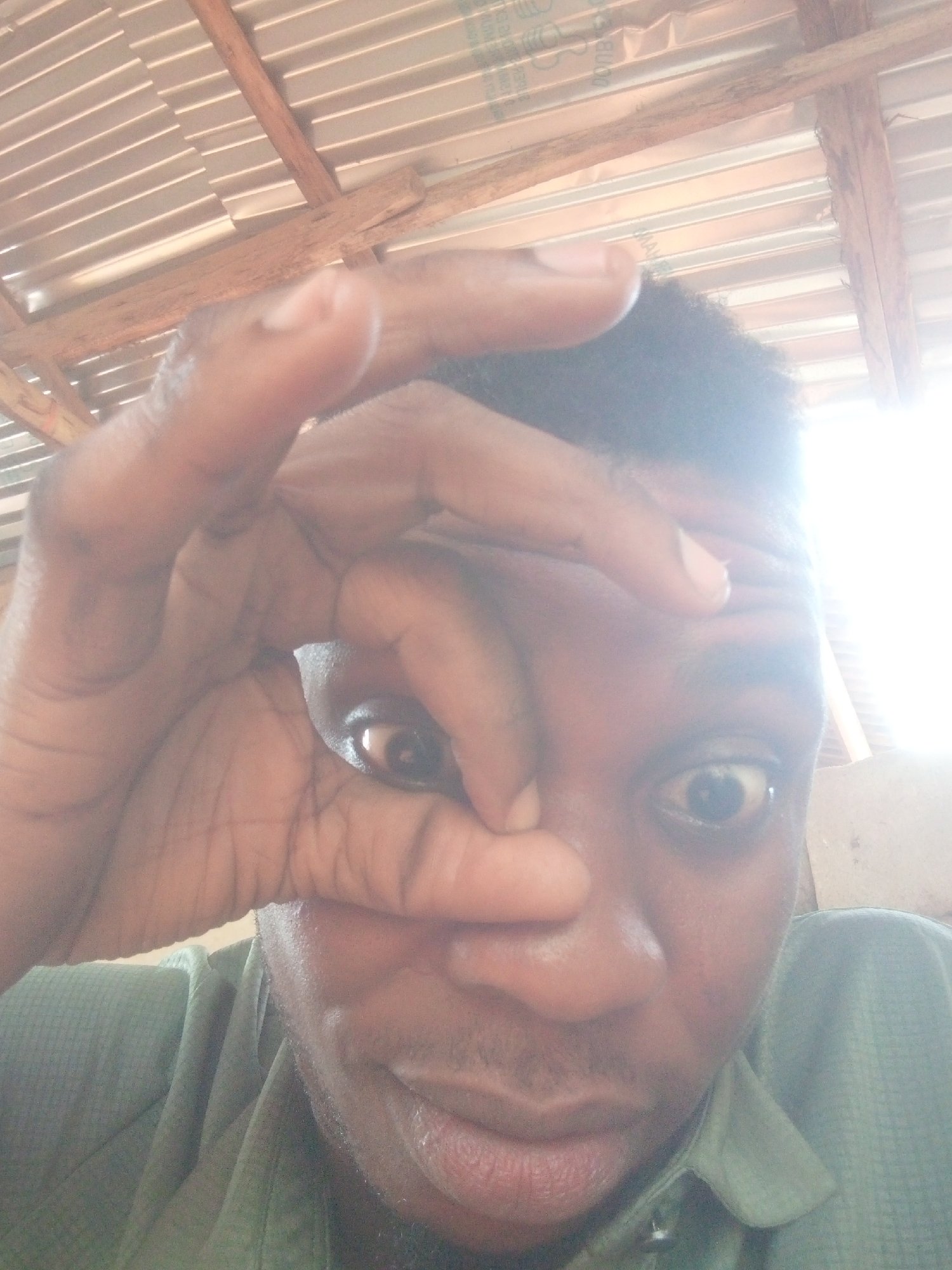 A close-up selfie shows a Black male, facing forward, with his hand positioned in a circular shape over one eye. His other eye is visible, looking towards the camera. He has short, dark hair and is wearing a light green collared shirt. The background includes what appears to be a metal ceiling with wooden beams. The setting appears to be indoors, but the city and specific location are not discernible from the image.