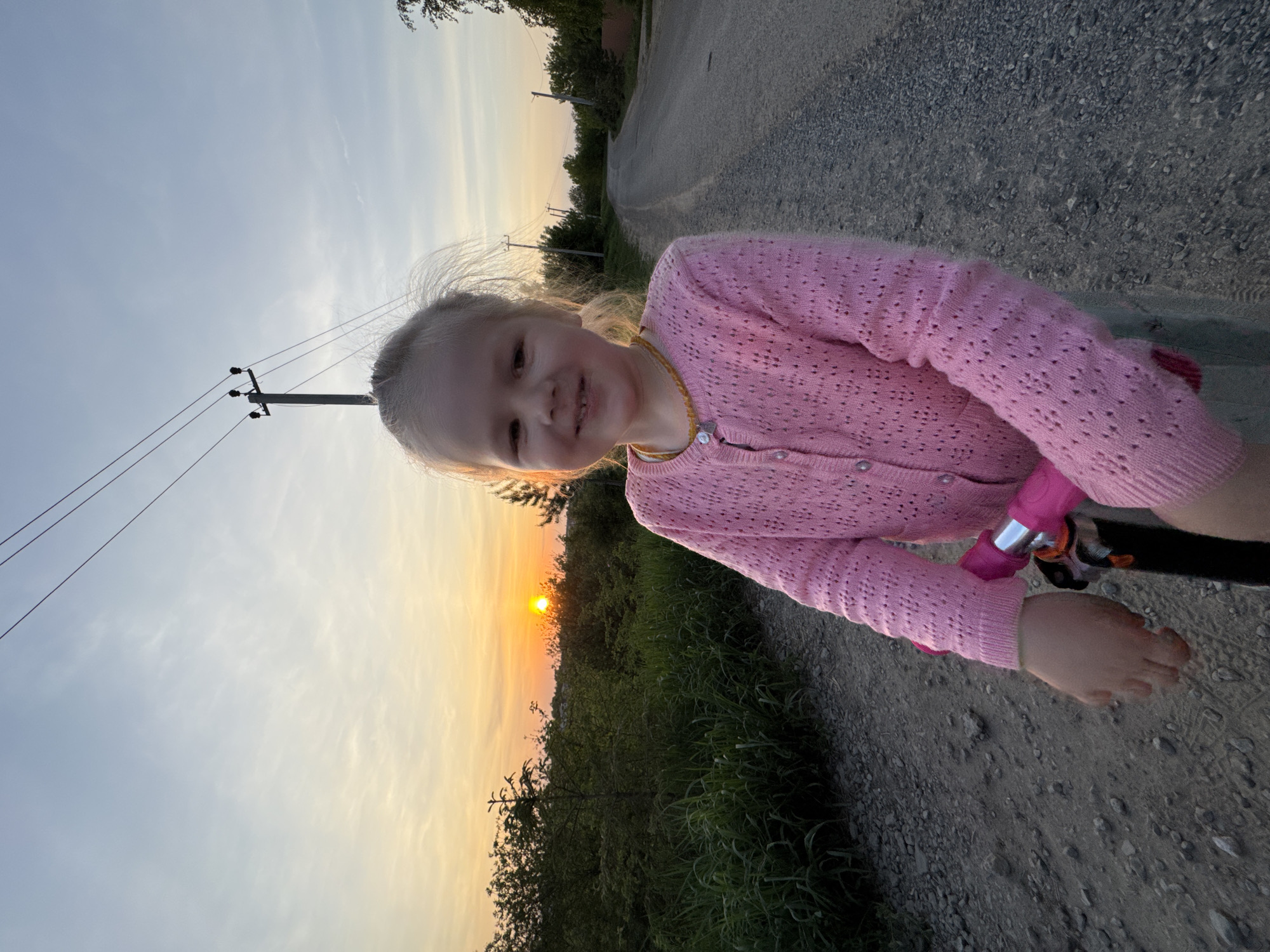 A young girl in a pink cardigan is riding a bicycle on a gravel road. The sun is setting in the background. There is a power pole on the left side of the image. The location is Горный, Russia.