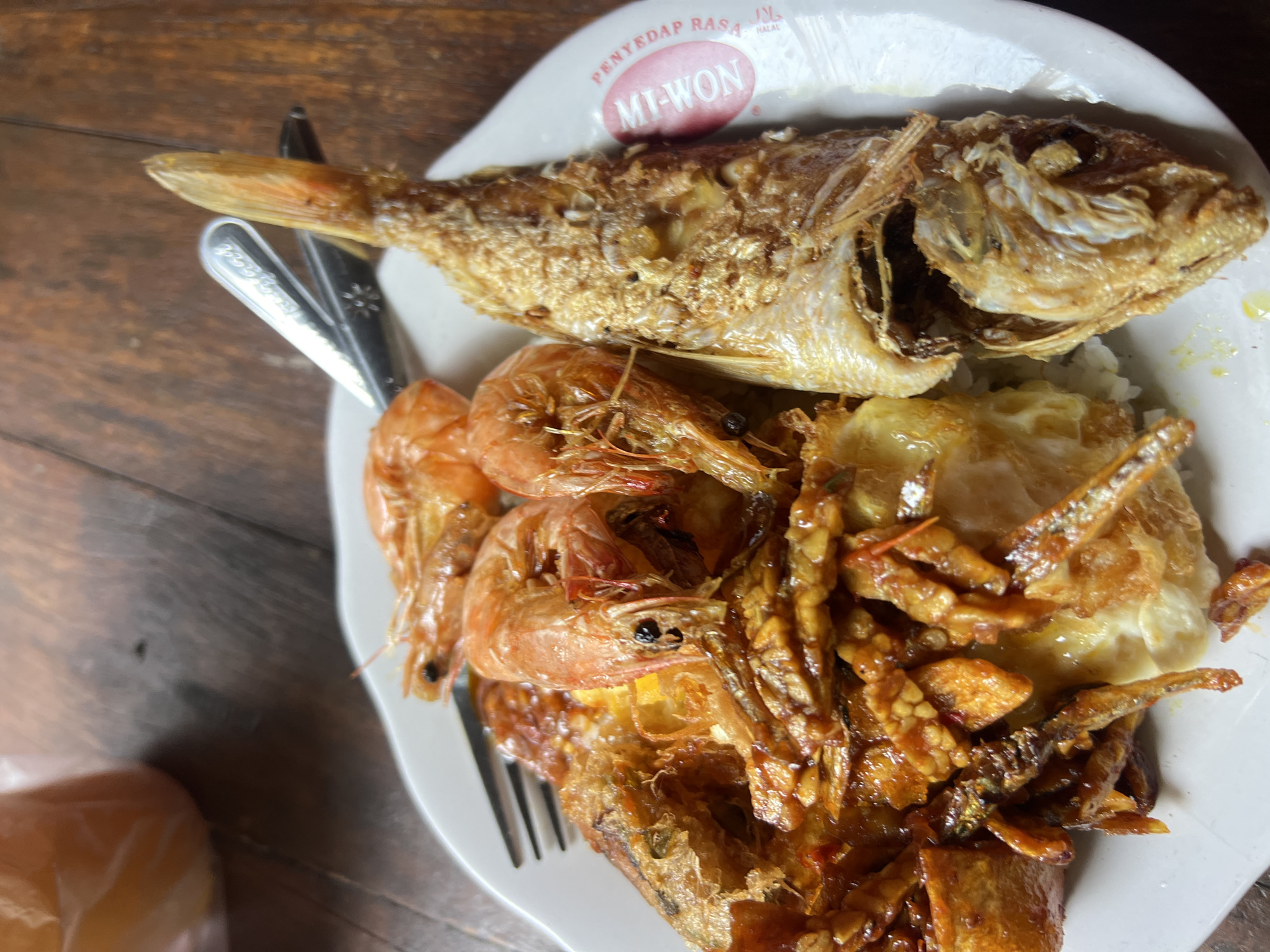 The image shows a close-up, overhead view of a white plate filled with food, set on a rustic wooden table. The meal consists of a whole fried fish, several large cooked shrimp, a fried egg with a glistening yolk, and a serving of small fried anchovies or similar small fish mixed with a reddish sauce, possibly chilies and other spices. The food appears to be a traditional Indonesian seafood dish. The plate has a logo on the right side that reads "Mt-Won" and "PENYEDAP RASA HALAL" in Indonesian, suggesting it is from a restaurant or food vendor in Indonesia. The background is blurred, showing more of the wooden table and a glimpse of a beverage in a clear plastic cup on the upper left. A fork and a spoon are partially visible, resting on the plate. The lighting suggests an outdoor or well-lit indoor setting. The food is presented in a casual, appetizing manner, indicative of a local eatery. The image does not contain any people or explicit actions or interactions. The scene is static and focuses solely on the food. The specific location in Kuta, Indonesia, cannot be determined from the visual information alone, beyond the general context provided by the Indonesian text on the plate.