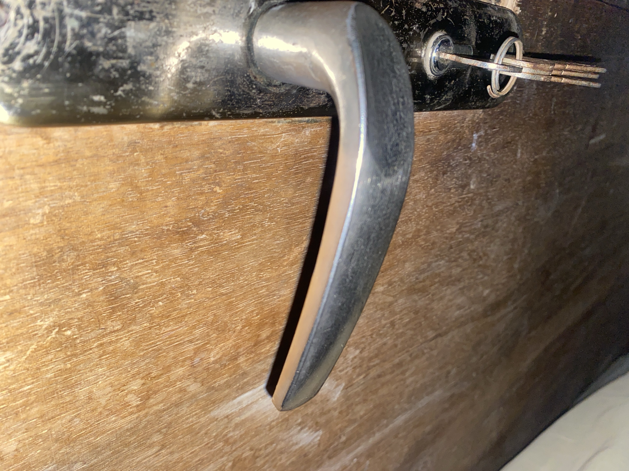 The image shows a close-up view of a weathered wooden door with a metal door handle and a set of keys inserted into the lock. The door handle is silver-toned and appears to be made of metal, showing some signs of wear and tear. A key is fully inserted into the lock mechanism, with its head and a keyring visible. The surrounding wooden surface of the door exhibits a visible grain pattern and appears to be aged. The lighting suggests an indoor environment, possibly a domestic setting. The scene is devoid of any individuals or immediate human activity.