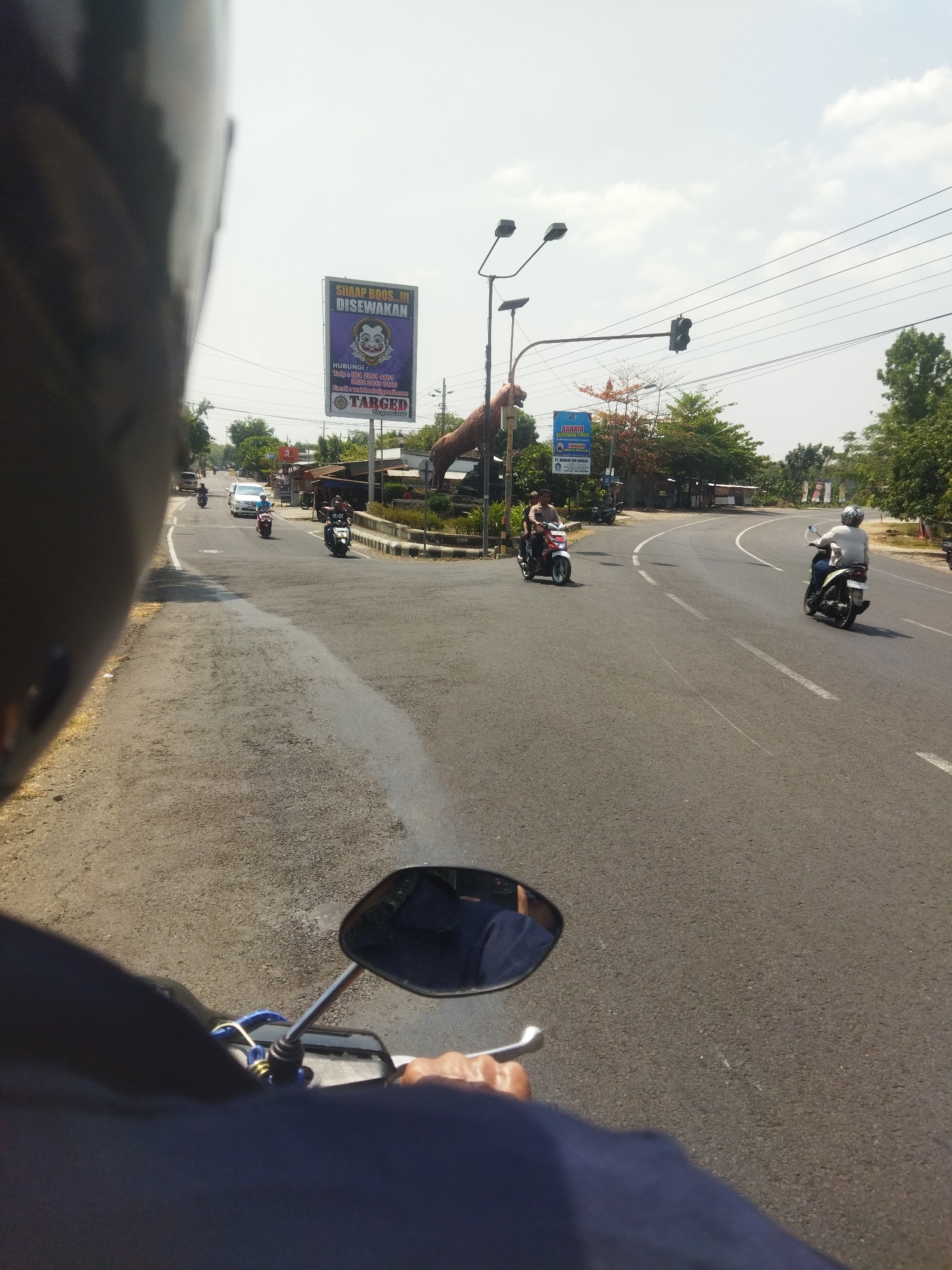 The image captures a first-person perspective of a motorcycle ride on a sunny day in Nambangan, Indonesia. The road is paved and appears to be a two-lane street with white lane markings. Several motorcycles are visible, some with riders and passengers, navigating the road. A white car is also present.
On the right side of the road, there are businesses with storefronts and signs, including a prominent billboard for "SHAAP ROOS...!!! DISEWAKAN". There are also trees and utility poles lining the street. A traffic light is visible in the distance. The sky is bright with some clouds.
The rider's helmet is partially visible in the foreground, and their hand is on the handlebars, suggesting the image was taken while the motorcycle was in motion. The overall scene depicts a typical urban environment in Indonesia, with the characteristic presence of motorcycles as a primary mode of transportation.