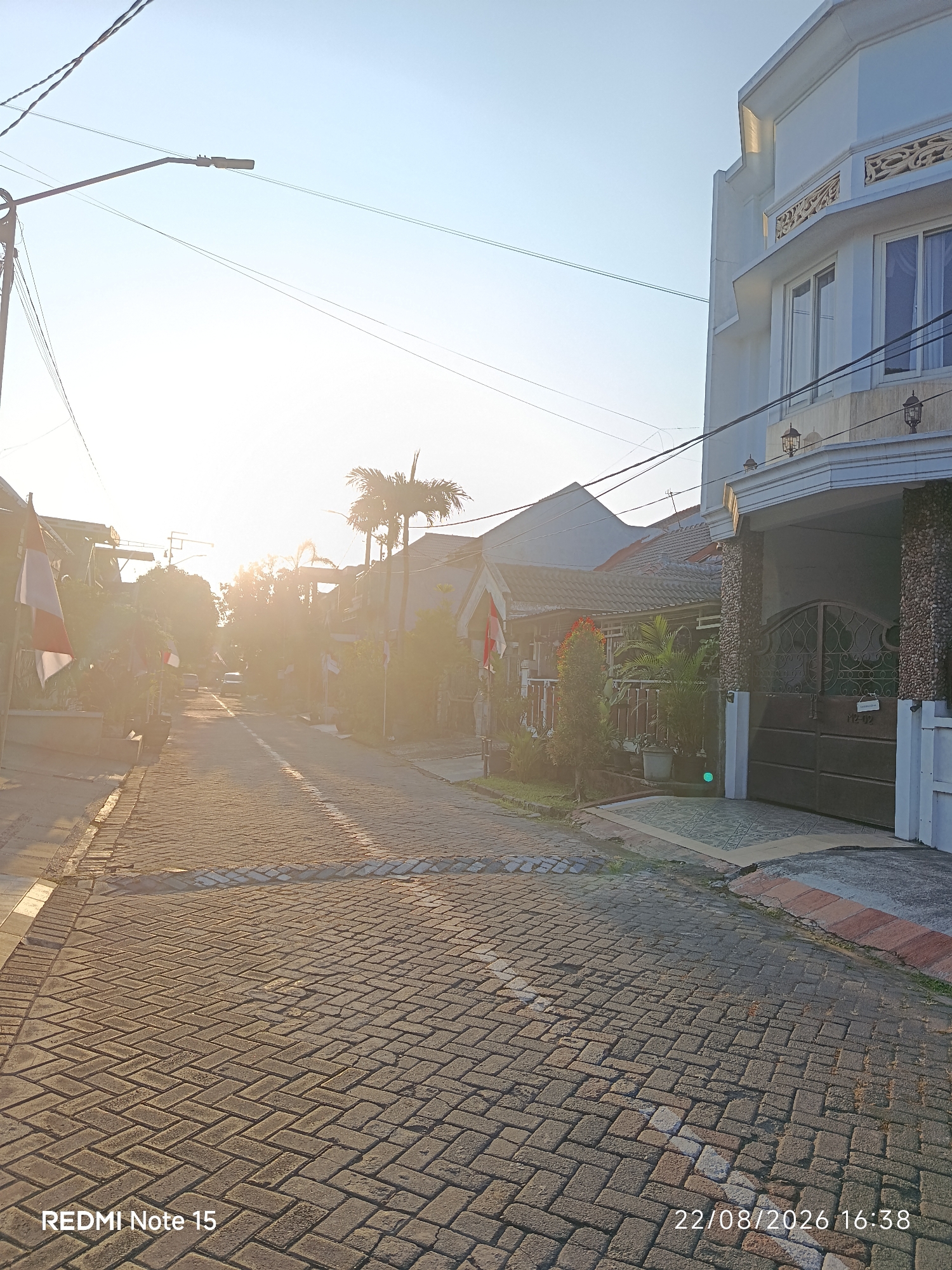 The image depicts a street scene in Keboansikep, Indonesia, at approximately 16:38 on August 22, 2026. The perspective is from a low angle, looking down a paved road lined with houses and vegetation. The sun is low in the sky, casting a warm glow and creating lens flare. On the left side of the road, flags of Indonesia are visible, mounted on poles. The road surface is paved with interlocking brick pavers laid in a herringbone pattern. Houses with tiled roofs and white facades are visible on both sides of the street. A large, modern white house with decorative stonework and a dark metal gate is prominent on the right side of the image. Overhead, electrical wires and a street lamp are suspended. The overall impression is a quiet residential street bathed in the warm light of late afternoon.