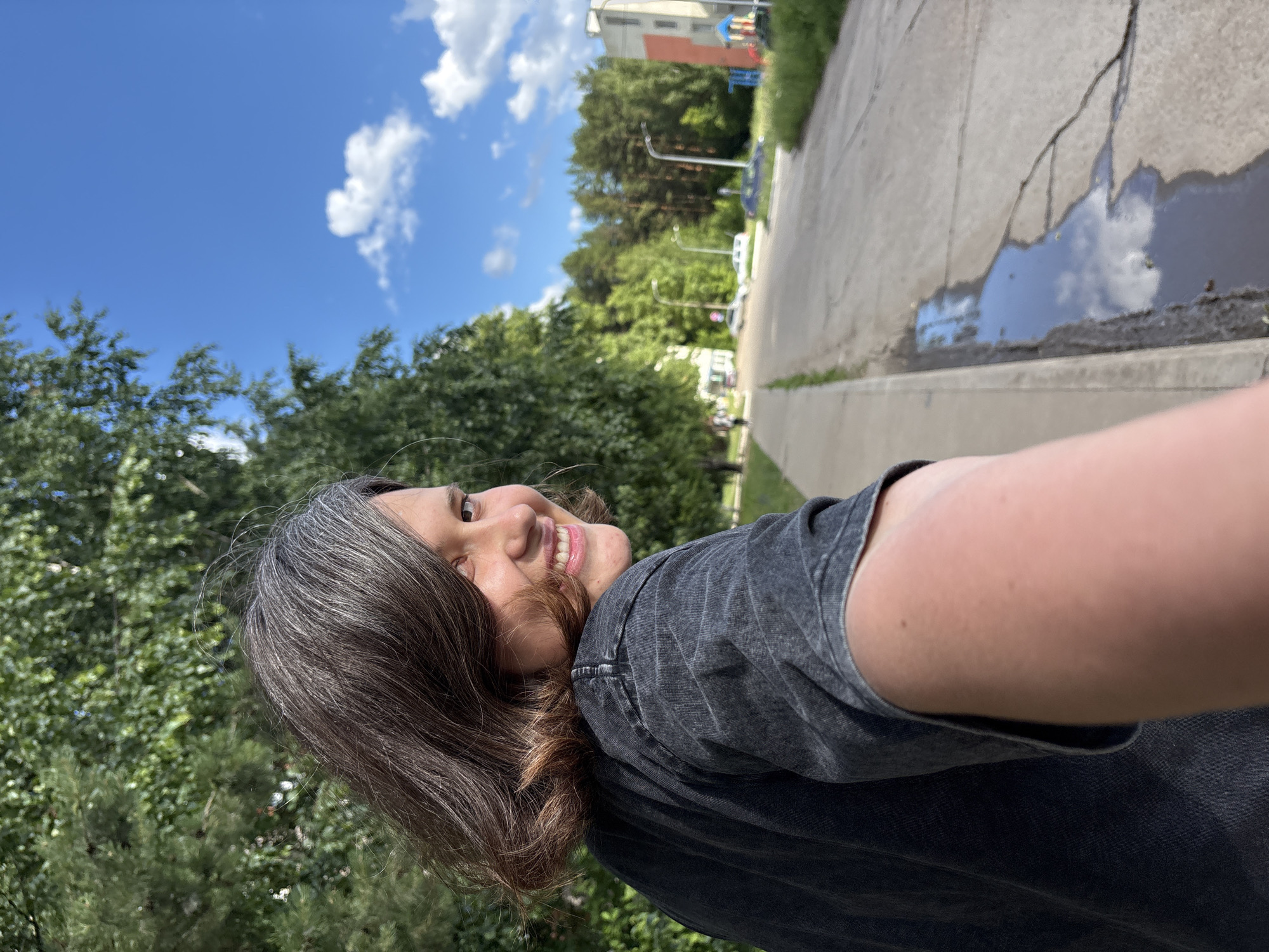 The media file is a portrait-style selfie taken outdoors on a sunny day. The subject, a person with brown hair showing some gray, is smiling and looking over their shoulder towards the camera. They are wearing a dark gray t-shirt.
The background consists of lush green trees on the left, and a paved walkway with a puddle reflecting the sky on the right. Beyond the walkway, a multi-story building with orange and white sections is visible, along with a playground in front of it. The sky is a vibrant blue with fluffy white clouds. The overall impression is of a pleasant, casual outdoor setting, possibly a park or residential area in Chaikovsky, Russia. The time of day appears to be midday given the bright sunlight and clear sky. The mood conveyed is cheerful and relaxed. There is no visible text in the image.