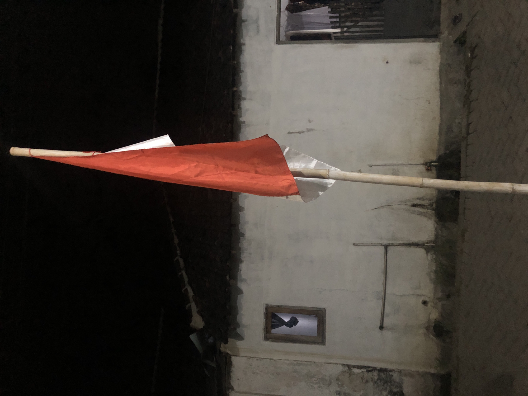 The image shows a red and white flag on a bamboo pole in front of a white building at night. The building has a dark roof and a small window with a dark curtain. To the right of the window is a doorway with a wooden gate. The ground in front of the building is paved with bricks. The scene is dimly lit, suggesting it is nighttime. The location is Tenggilis Rejo, Indonesia.