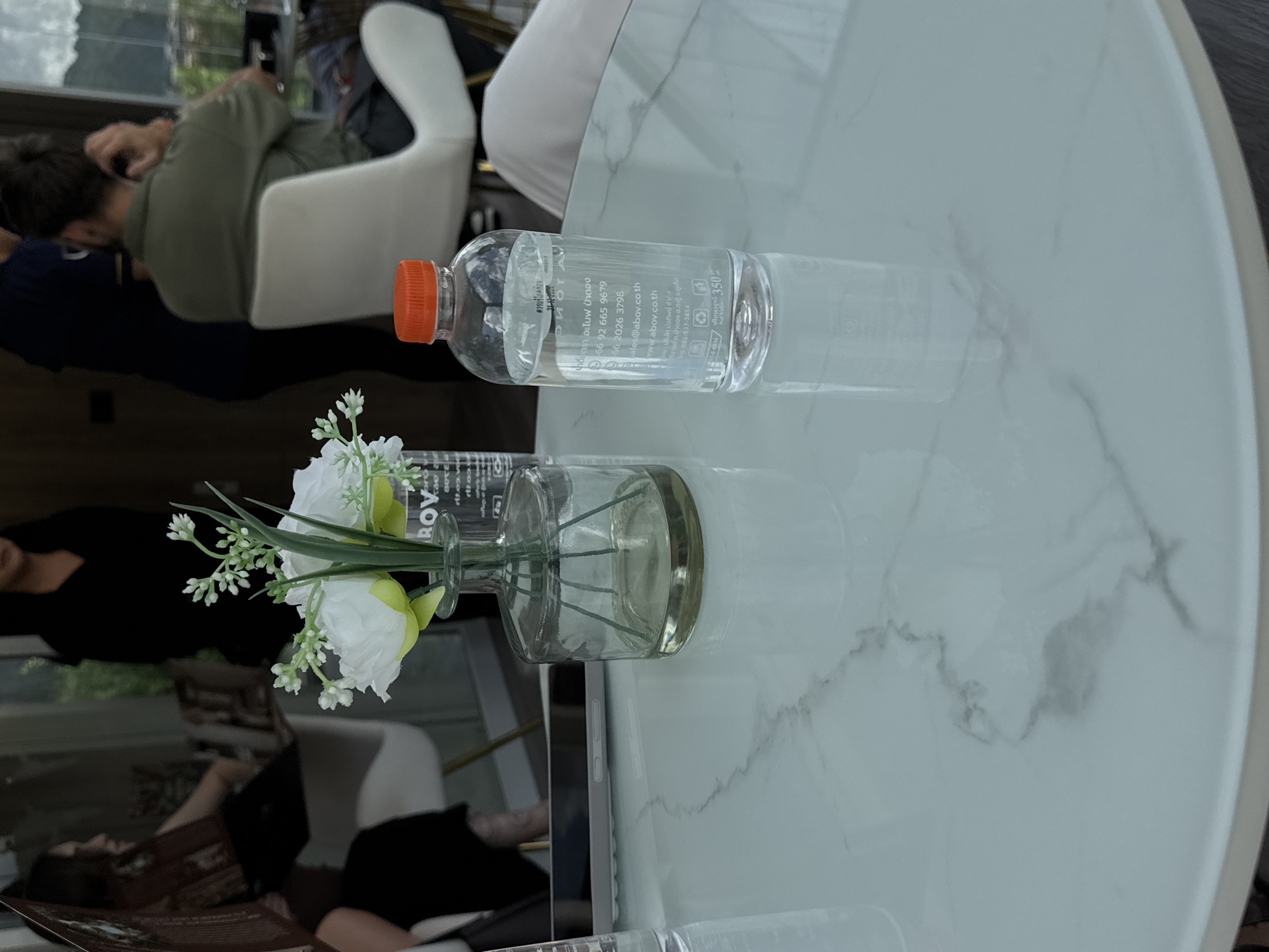 The image depicts a marble-patterned table in what appears to be an indoor cafe or restaurant setting in Patong, Thailand. On the table, a glass vase holds a small arrangement of white and green artificial flowers. Next to the vase is a clear plastic bottle of water with an orange cap, containing liquid and bearing a label with text in Thai and English. The bottle indicates a net volume of 350 ml. In the blurred background, several individuals are visible. One person, wearing a dark top, stands near a window. Another person, in a green shirt, is seated, with their back to the camera, appearing to be having their hair styled. Another figure, in a dark outfit, is partially visible to the left. The overall ambiance suggests a relaxed, possibly commercial, environment.