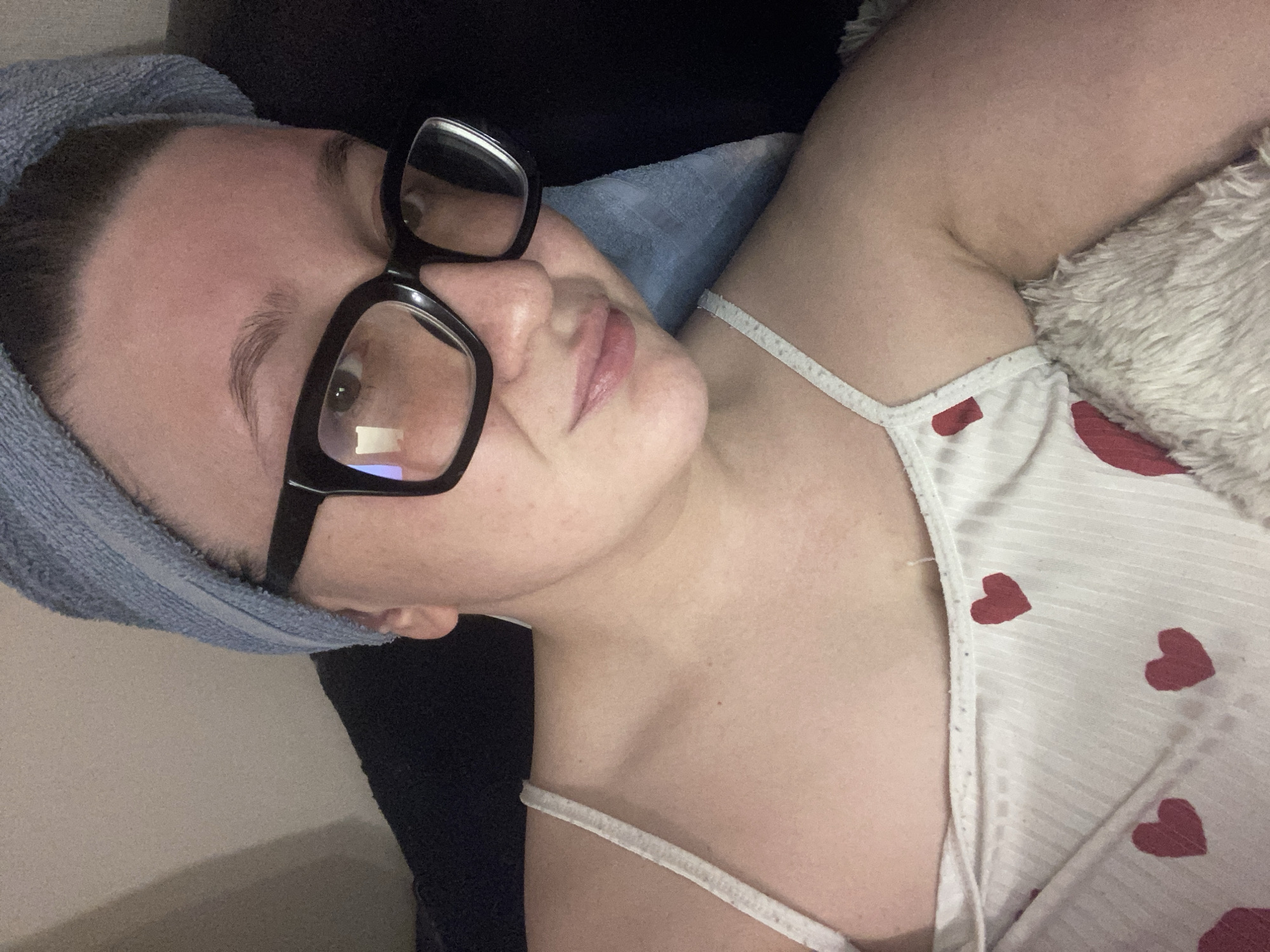 A young person with fair skin, wearing black-rimmed glasses and a light blue towel wrapped around their head, is lying down. They are wearing a white top with red heart patterns and thin straps. A fluffy, light-colored object is situated near their shoulder. The person is positioned against a dark background. No details indicate the location is in the city of Chaikovsky, Russia.