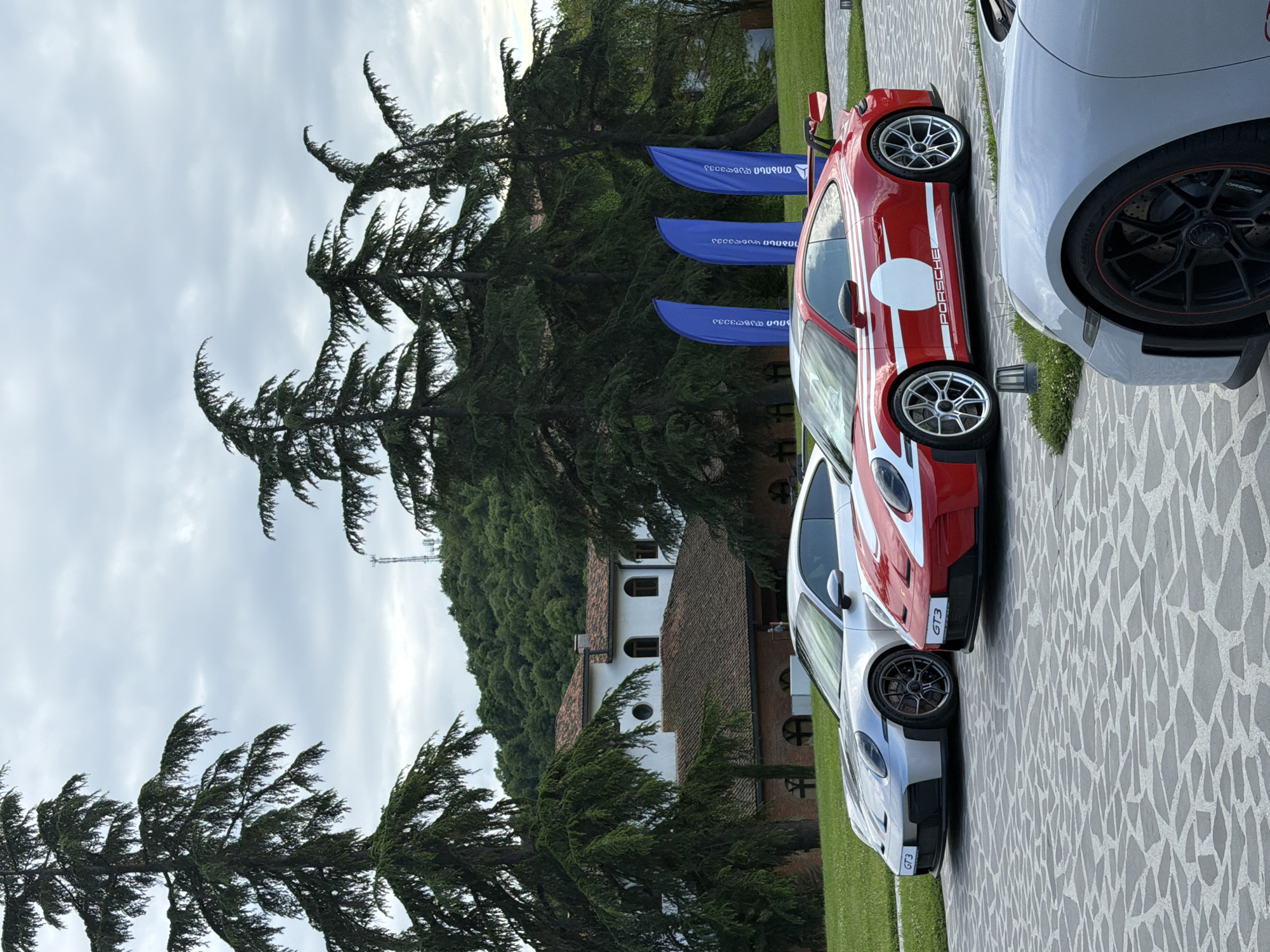The image depicts a scene in Napareuli, Georgia, showing three Porsche GT3 sports cars parked on a patterned stone surface. The cars are positioned side-by-side, with colors red, white, and gray. Blue flags are visible behind the red car. Trees and a building are in the background, under a cloudy sky. A cell tower can be seen among the trees in the background.