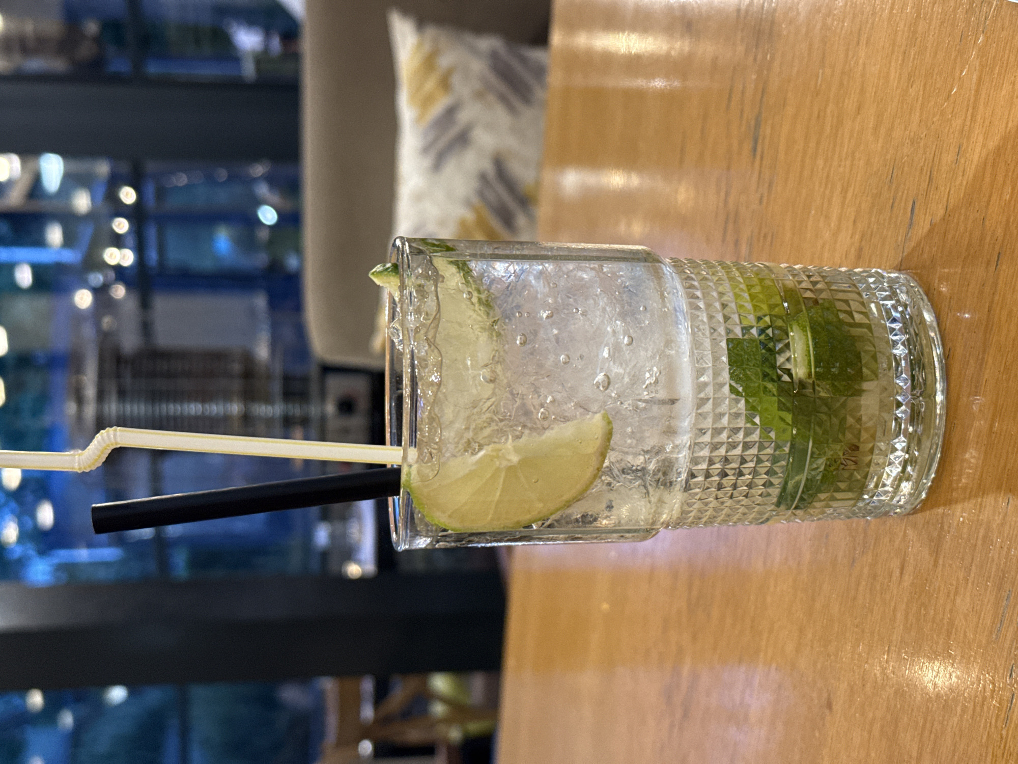 The image shows a glass of a clear drink, likely a cocktail, filled with ice and lime slices, placed on a wooden table. Two straws, one black and one yellow, are in the drink. The glass is embossed with a diamond pattern. The background is blurred and suggests a cafe or bar setting with lights visible. In the background, there is a pattern pillow and a grey back. The location is Pleven, Bulgaria.