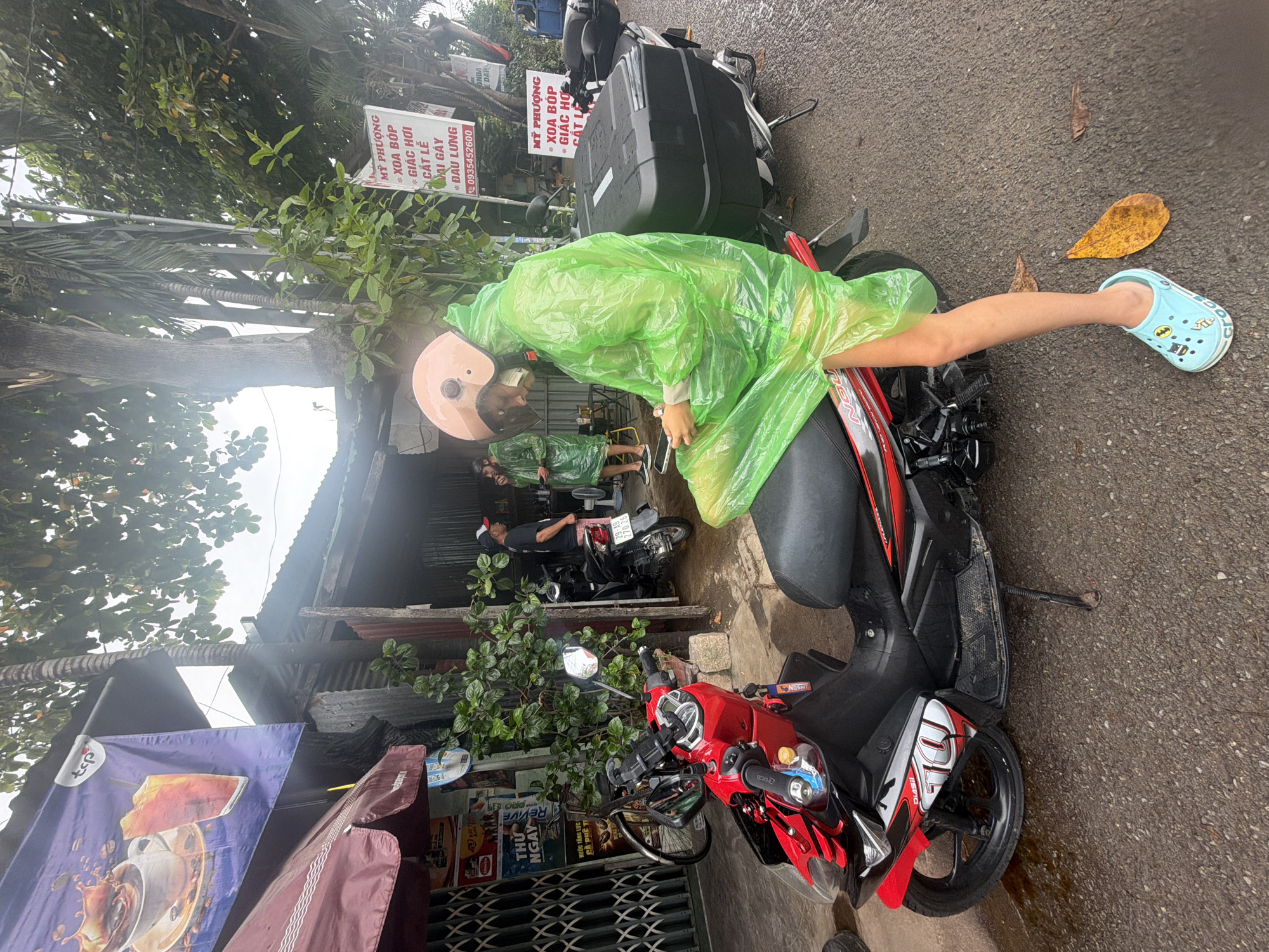 A person wearing a bright green plastic poncho and helmet sits on a red and black motorcycle on a paved street in Nha Trang, Vietnam. The person is looking down at their phone. A second person, also wearing a green poncho, stands in the background. The street appears wet, suggesting it has been raining. There are several shops and signs in Vietnamese along the street, including advertisements for coffee and other products. A large black storage box is attached to the back of the motorcycle. Fallen leaves are scattered on the ground.
