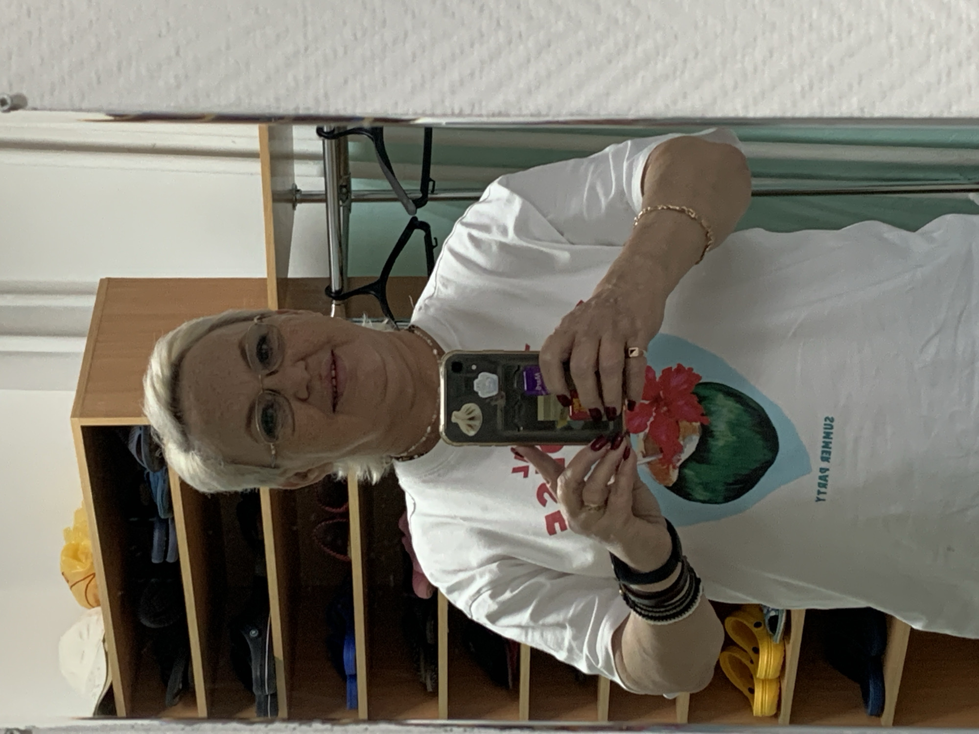 A Caucasian woman with short blonde hair and glasses is taking a selfie in a mirror. She is wearing a white t-shirt with a graphic of a coconut with flowers on it and the text "SUMMER PARTY" printed in reverse. She has dark red nail polish on and is wearing several bracelets on her left wrist and a gold chain bracelet on her right wrist. She is holding a smartphone in her right hand. Behind her, a wooden shelf unit is visible, filled with various shoes. To the right of the shelf unit, a clothing rack with hangers and a green curtain can be seen. The background suggests an indoor setting, likely a home or personal space. This image was taken in Chaikovsky, Russia.
