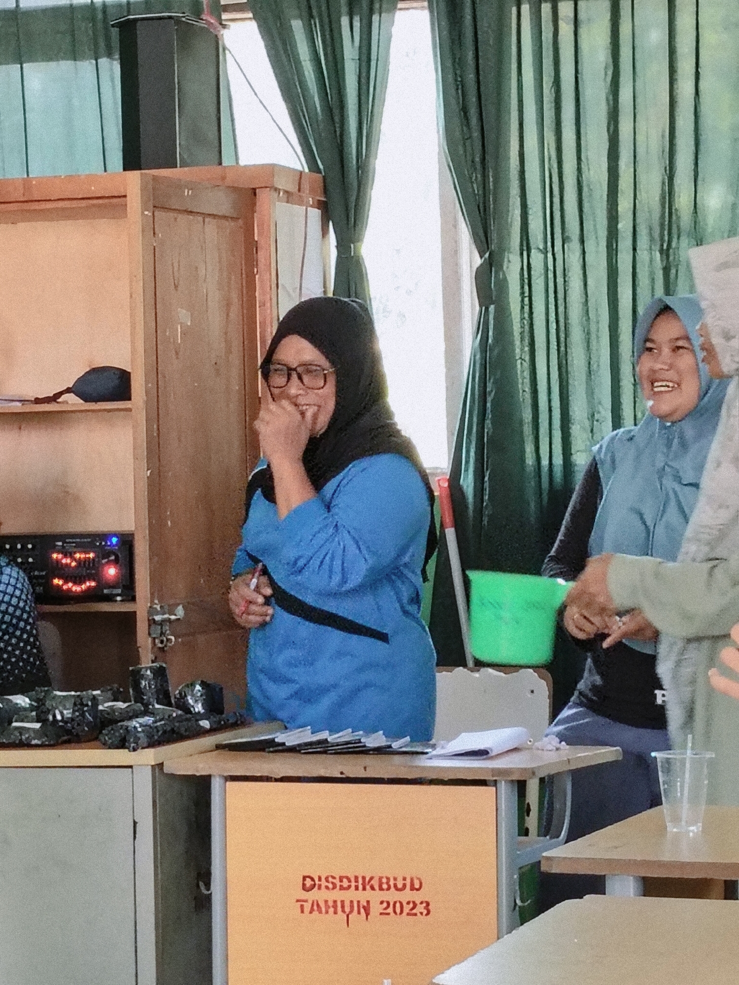 In what appears to be an indoor event space, possibly a classroom or community hall in Lori, Indonesia, three women are visible. The woman in the foreground, wearing glasses and a black hijab, is dressed in a blue top and is laughing, holding a pen in her right hand. To her right, another woman in a light blue hijab is also smiling broadly. A third woman, partially visible in a light gray hijab, stands on the far right. Behind the women, a shelf unit with a speaker on top is present, along with green curtains and natural light filtering in from a window. In front of the women, a table displays various items, including what look like black fabric samples, a small green bucket, and a clear plastic cup. The overall atmosphere suggests a casual, perhaps educational or celebratory gathering.