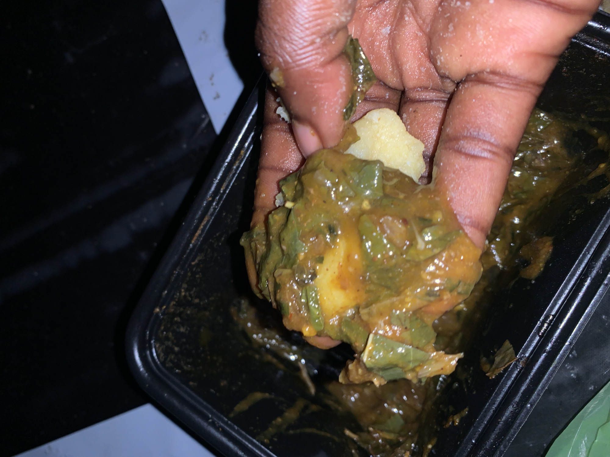 A hand is holding a portion of a green stew with chunks of starchy food, likely fufu, in a black plastic container. The stew appears to be of a moderate consistency with visible pieces of leafy greens and some orange-colored spices or peppers. The hand is dark-skinned and some white residue, possibly salt or flour, is visible on the fingers. The background is dark and out of focus, with hints of a light-colored surface. This scene is located in Rafin Par, Nigeria.