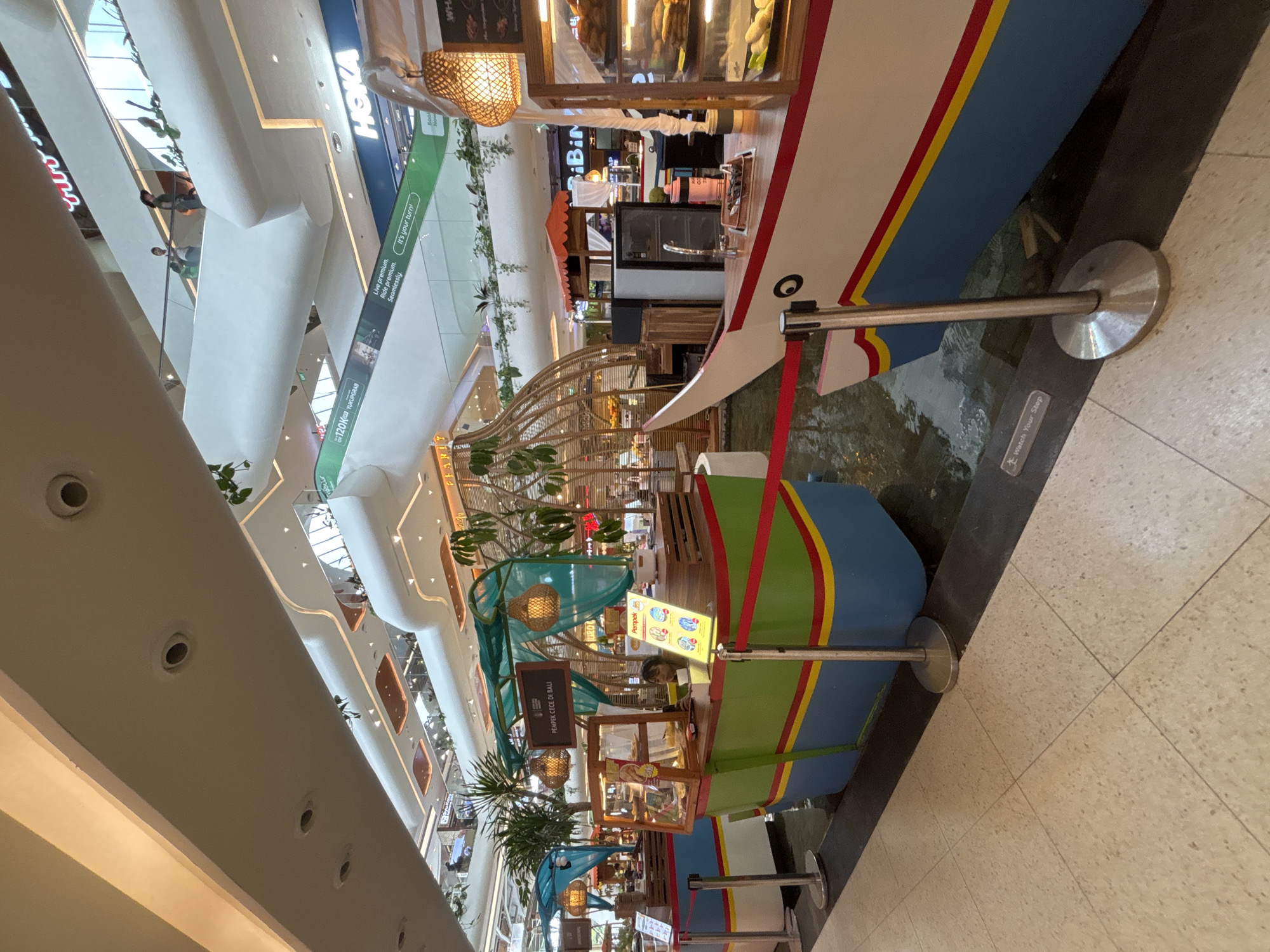 The image shows a food court in Denpasar, Indonesia. Several food stalls are designed to resemble boats. The stalls are brightly colored with blue, green, and red paint. Signs advertise various items, including "PEMPEK CECE DI BALI" and "Pempek." A woman is visible behind one of the stalls. There are stanchions with silver posts and flat circular bases. The floor is light tan tiles. The ceiling is high with a modern design, including spotlights. In the distance, there are people walking and shopping. The lighting is bright.