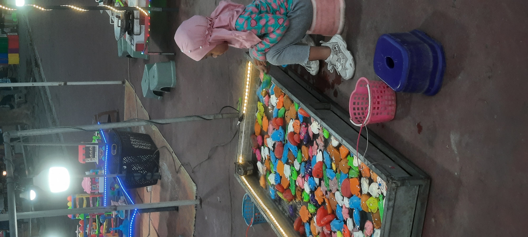 A young girl, wearing a pink hijab and a long-sleeved shirt with a heart pattern, sits on a pink stool and plays a fishing game at a carnival stall in Kanigoro, Indonesia. The game consists of a large rectangular tank filled with water and numerous colorful plastic fish. The girl is reaching into the tank with a fishing rod. A pink basket is placed near the tank. In the background, there are carnival games and stalls with bright lights and colorful decorations. The ground is paved with reddish-brown concrete.