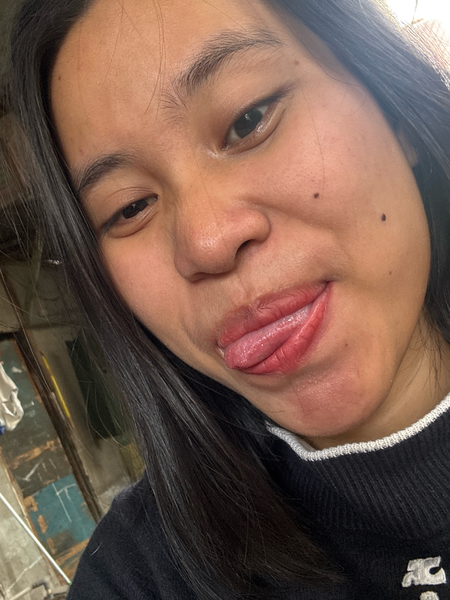 A close-up selfie features a young woman in East Jakarta, Indonesia, with her face tilted slightly to her right. She has dark hair and is wearing a black sweater with a white trim collar. Her lips are glossy and slightly parted, with her tongue extended, showing a glistening texture. Two dark moles are visible on her right cheek. The background is blurred and appears to be an indoor setting with wooden and possibly metal elements, suggesting a utilitarian or workshop environment.