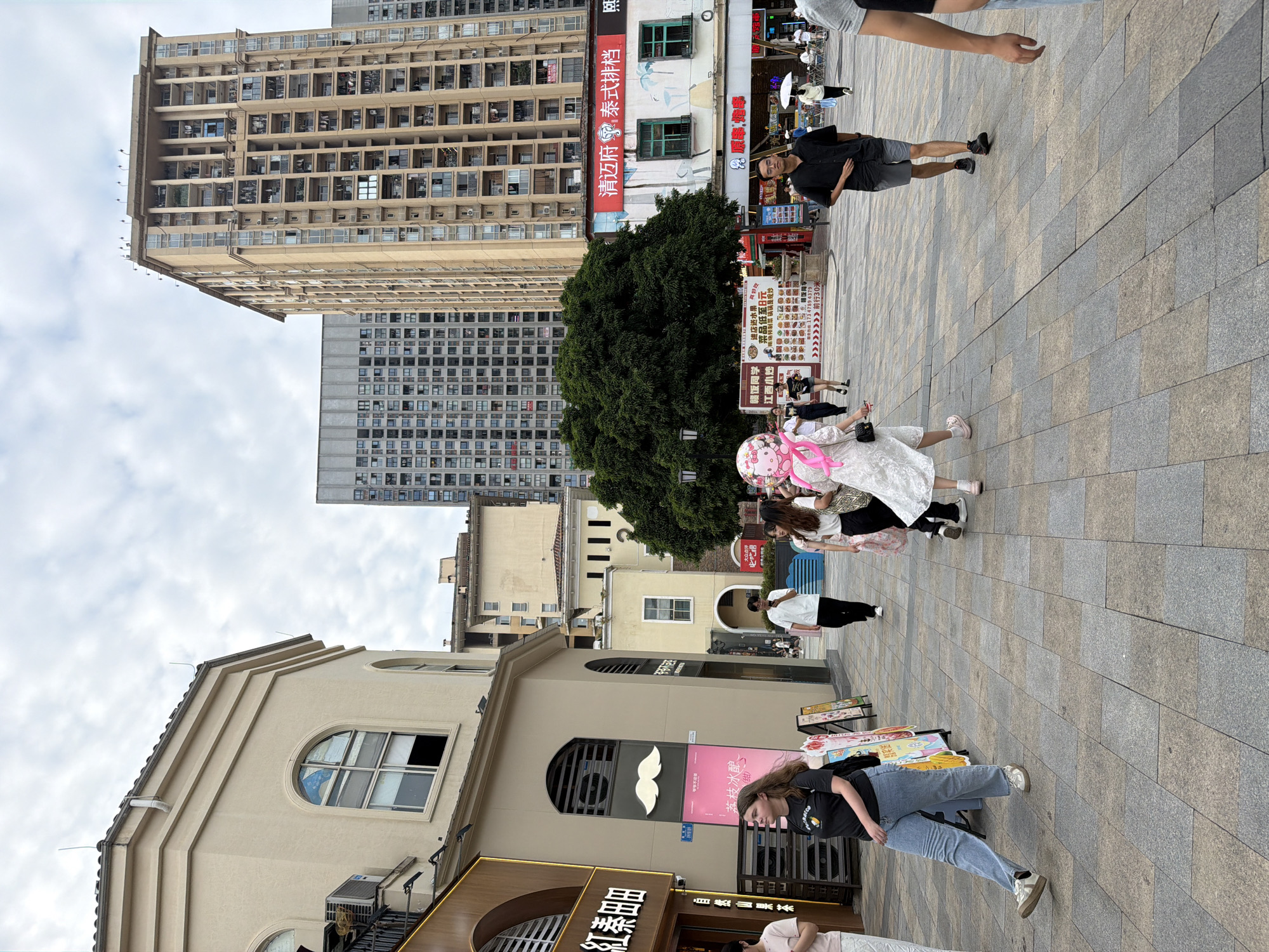 The image depicts an outdoor urban scene in the Chongqing High-tech Zone, China, characterized by a pedestrian walkway paved with interlocking stone tiles. In the foreground, several individuals are walking. A young woman with long, light brown hair, wearing a black t-shirt and blue jeans, is moving from left to right, her left leg extended. To her right, a woman in a white dress with a pink bow accessory on her head walks towards the camera, holding a large pink balloon shaped like Hello Kitty. Behind her, other people are also walking, some holding umbrellas.
The background reveals a mix of architectural styles. To the left, a light-colored building with arched windows and an air conditioning unit mounted on its exterior is visible. Further back, several tall, multi-story residential buildings dominate the skyline, showcasing a dense urban environment with numerous balconies and windows. A large, dense green tree stands prominently in the middle ground, providing a contrast to the surrounding concrete structures. Red signage with Chinese characters is present on some of the buildings, indicating commercial establishments. The sky is overcast with scattered clouds. The overall impression is of a busy public space within a modern Chinese city.