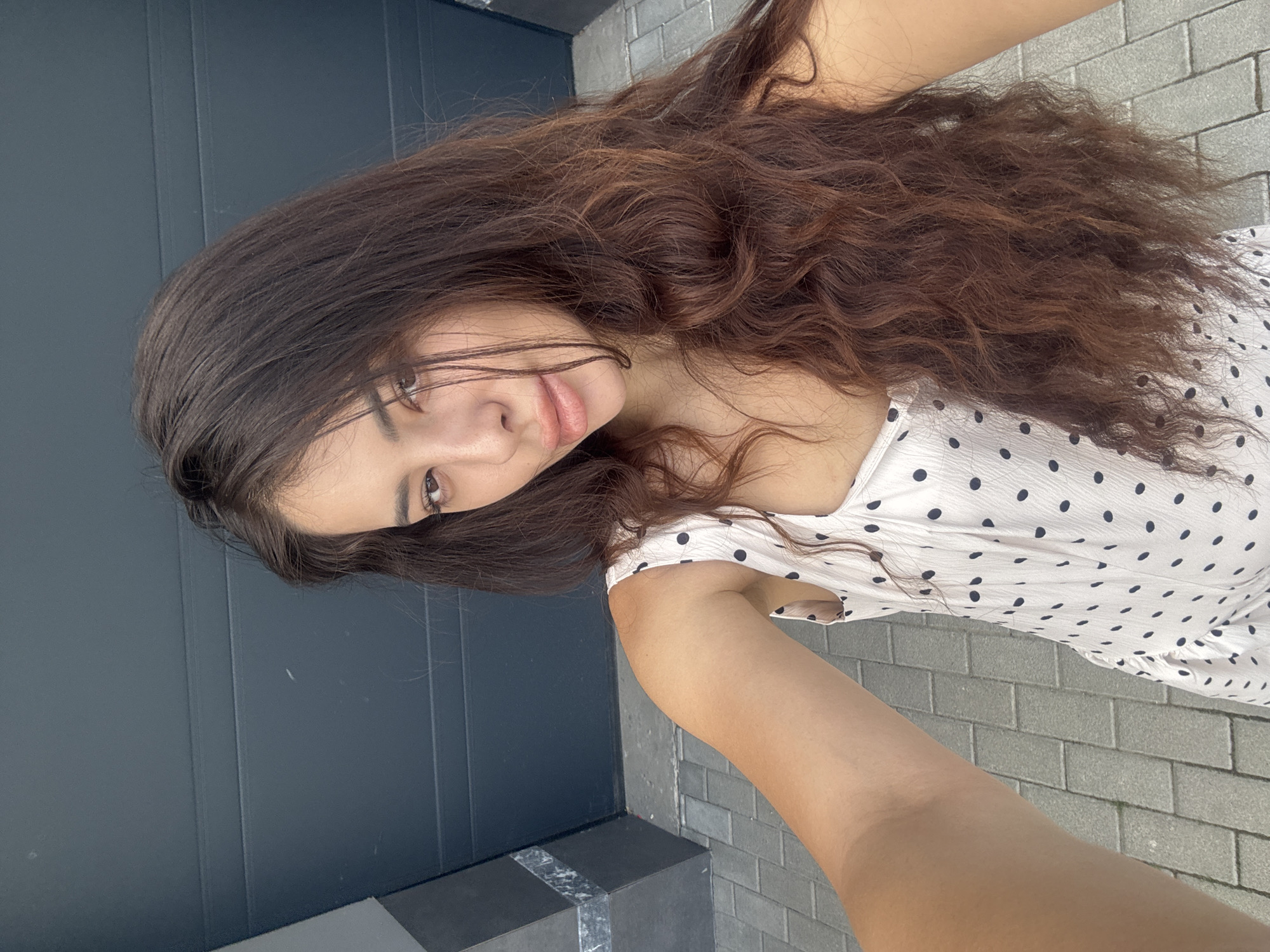 A young woman poses for a selfie outdoors. She has long, wavy dark brown hair and is wearing a white dress with black polka dots. The background consists of a dark gray door and a gray brick wall. No further detail allows to determine if the image was taken in Plovdiv, Bulgaria.