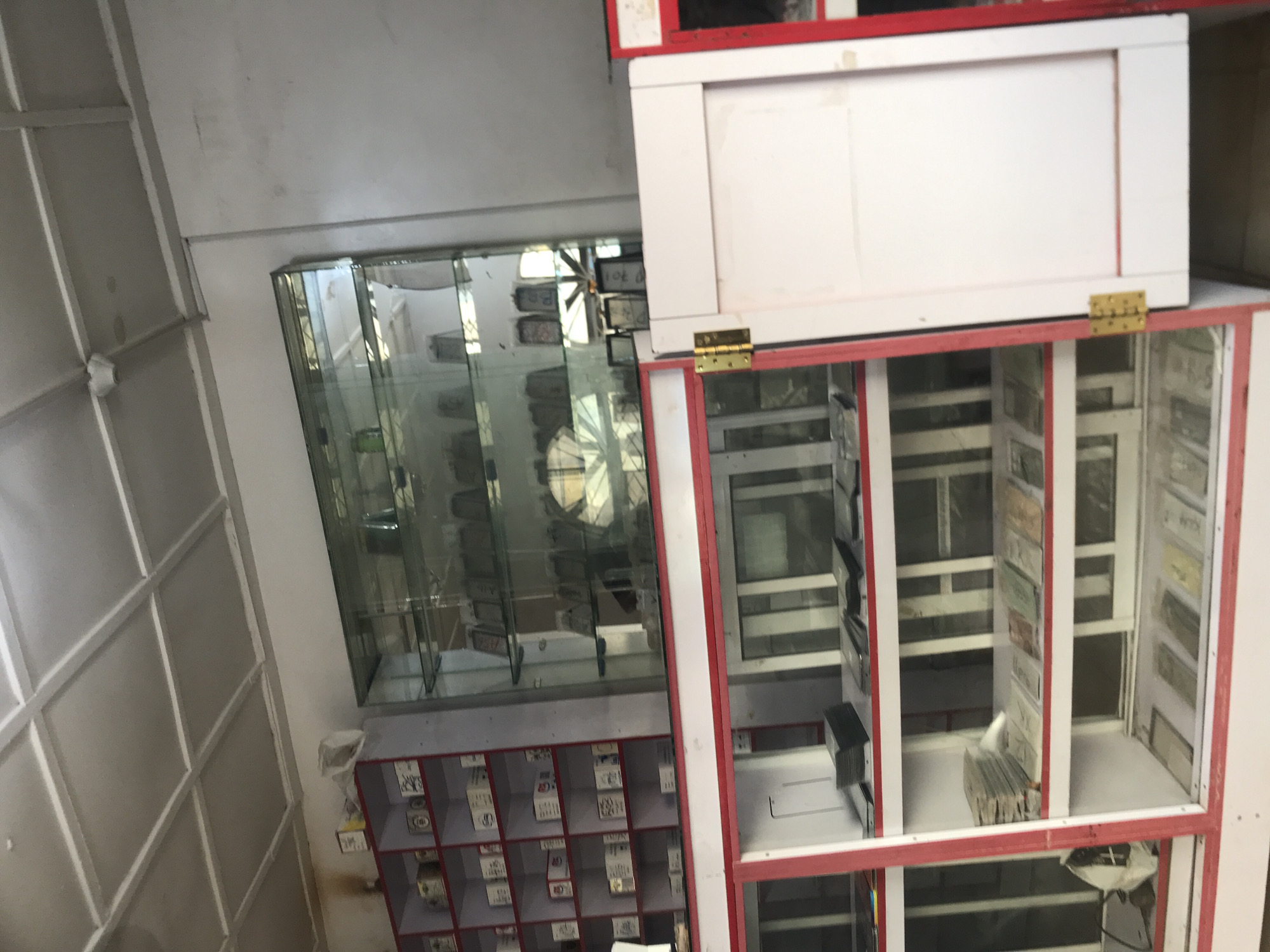 The image depicts the interior of a shop, likely in Jos, Nigeria, filled with various items displayed on shelves and in glass cabinets. On the left, a red and white shelf unit is packed with small boxes, some with handwritten labels. Adjacent to this is a tall glass display cabinet filled with numerous small items, possibly collectibles or electronics, arranged on multiple shelves. Below the glass cabinet, a red-trimmed display counter contains rows of what appear to be flat items like photos, cards, or small packets, also organized on shelves. A white door with a brass hinge is open on the right side of the counter. The ceiling is a grid of white panels. The overall impression is that of a retail space stocked with a variety of goods.