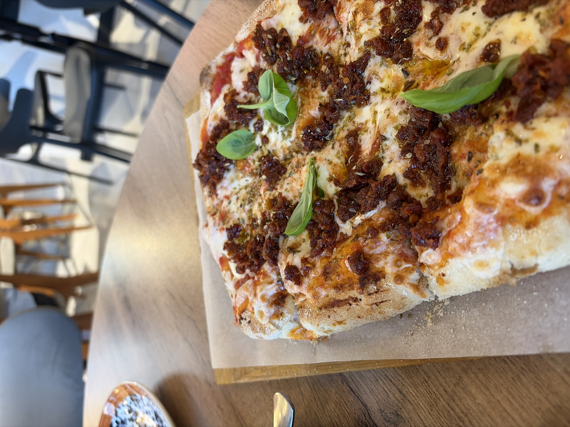 A high-angle shot displays a square pizza on a wooden serving board placed on a wooden table, likely in a restaurant. The pizza features a thick crust, a tomato sauce base, melted cheese, and a generous topping of what appears to be dried tomatoes and herbs. Fresh basil leaves garnish the pizza. A partial view of a chair, a metal utensil, and a small plate appear in the blurred background. The setting is indoors, likely a restaurant or cafe. The absence of people and contextual clues makes it impossible to determine if the location is in Izhevsk, Russia, or anywhere else in the world.