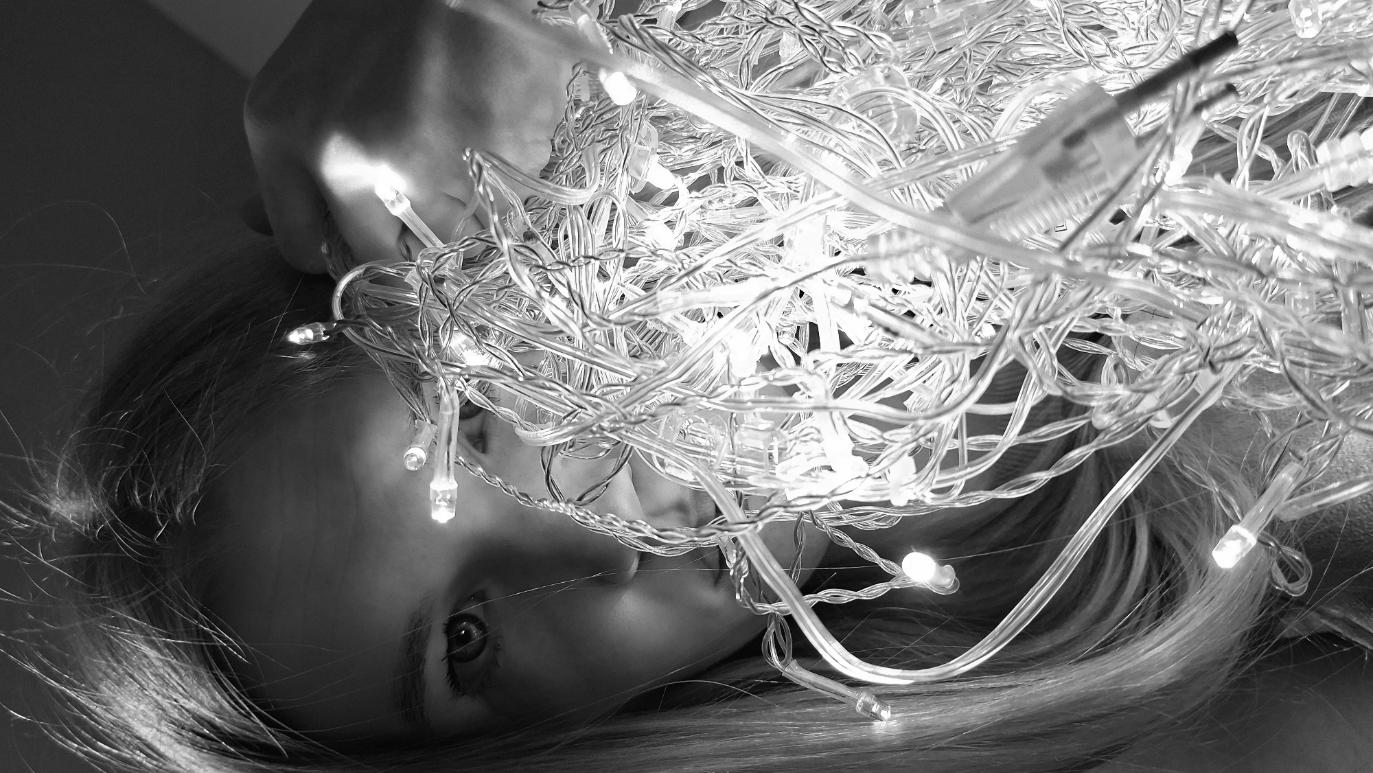 A black and white image captures a close-up of a young person, likely female, partially covered or lying beneath a dense tangle of illuminated string lights. Her face is seen from a slightly angled perspective, with one eye clearly visible and looking towards the viewer. Long, light-colored hair spills out from behind her head. A hand is visible in the upper left, seemingly resting near the mass of lights.
The primary objects in the scene are the string lights themselves, featuring numerous small, bright LED-style bulbs and clear wires, which create a chaotic but luminous pattern across the frame, especially prominent in the upper and central areas. A power connector for the lights is also visible in the upper right quadrant. The bright lights cast highlights on the person's skin and hair, and also create strong contrasts and shadows throughout the image.
The setting appears to be an indoor, low-light environment, which accentuates the glowing effect of the lights. The background is dark and indistinct. No specific activity is clearly depicted other than the person's presence amidst the lights, possibly posing or interacting with them. The mood is ethereal and atmospheric, enhanced by the monochrome filter. There is no visible text in the image.