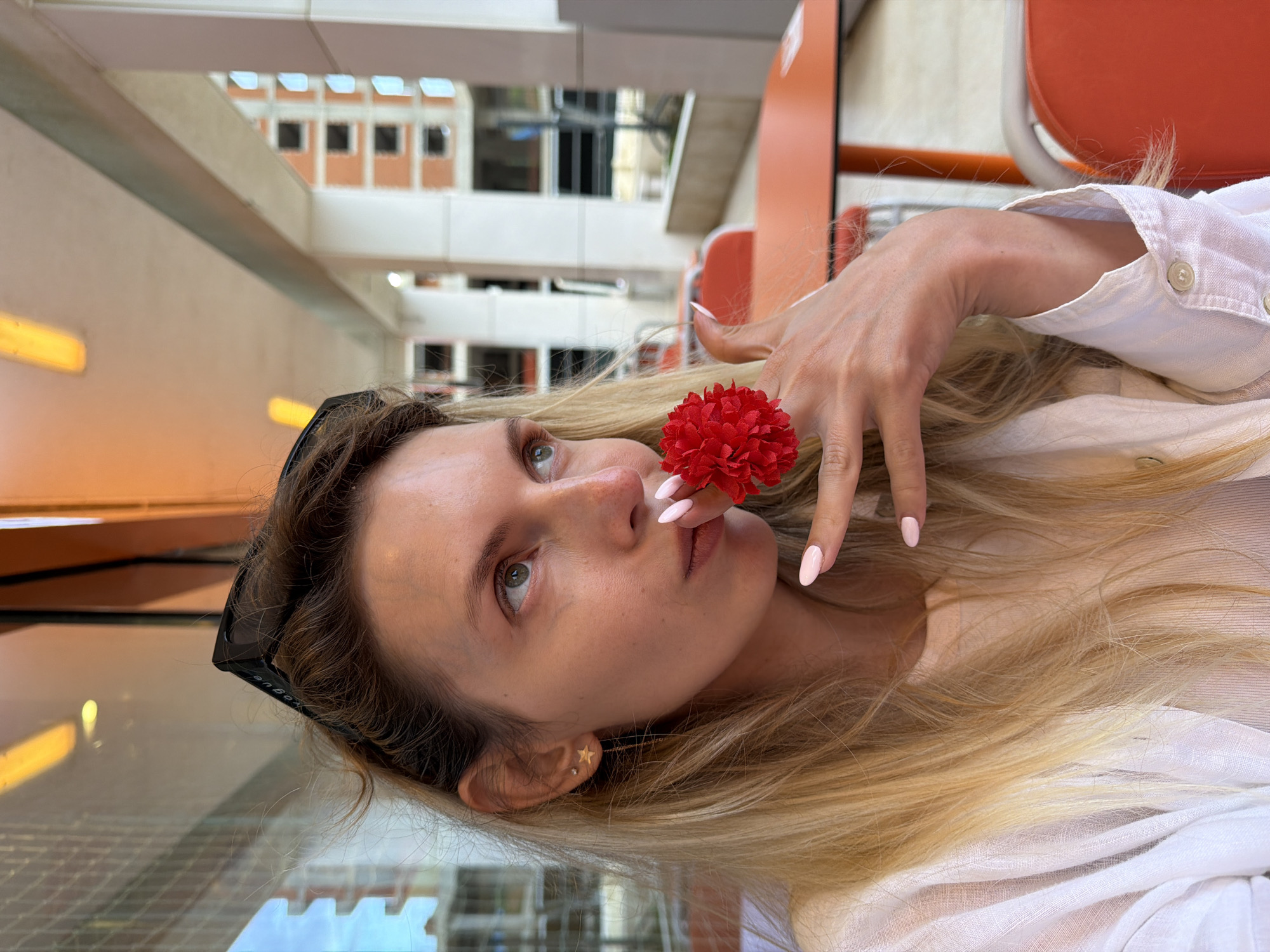 A woman with long blonde hair is holding a red flower near her mouth, appearing to be indoors, possibly on a train or ferry. She's wearing a white button-up shirt and has sunglasses tucked into her hair. The background features a building with a repetitive window pattern, suggesting an institutional or commercial structure, and there are orange elements which may be seats. There are no obvious signs indicating that this photograph was taken in Budva, Montenegro. Further information would be needed to confirm the location.