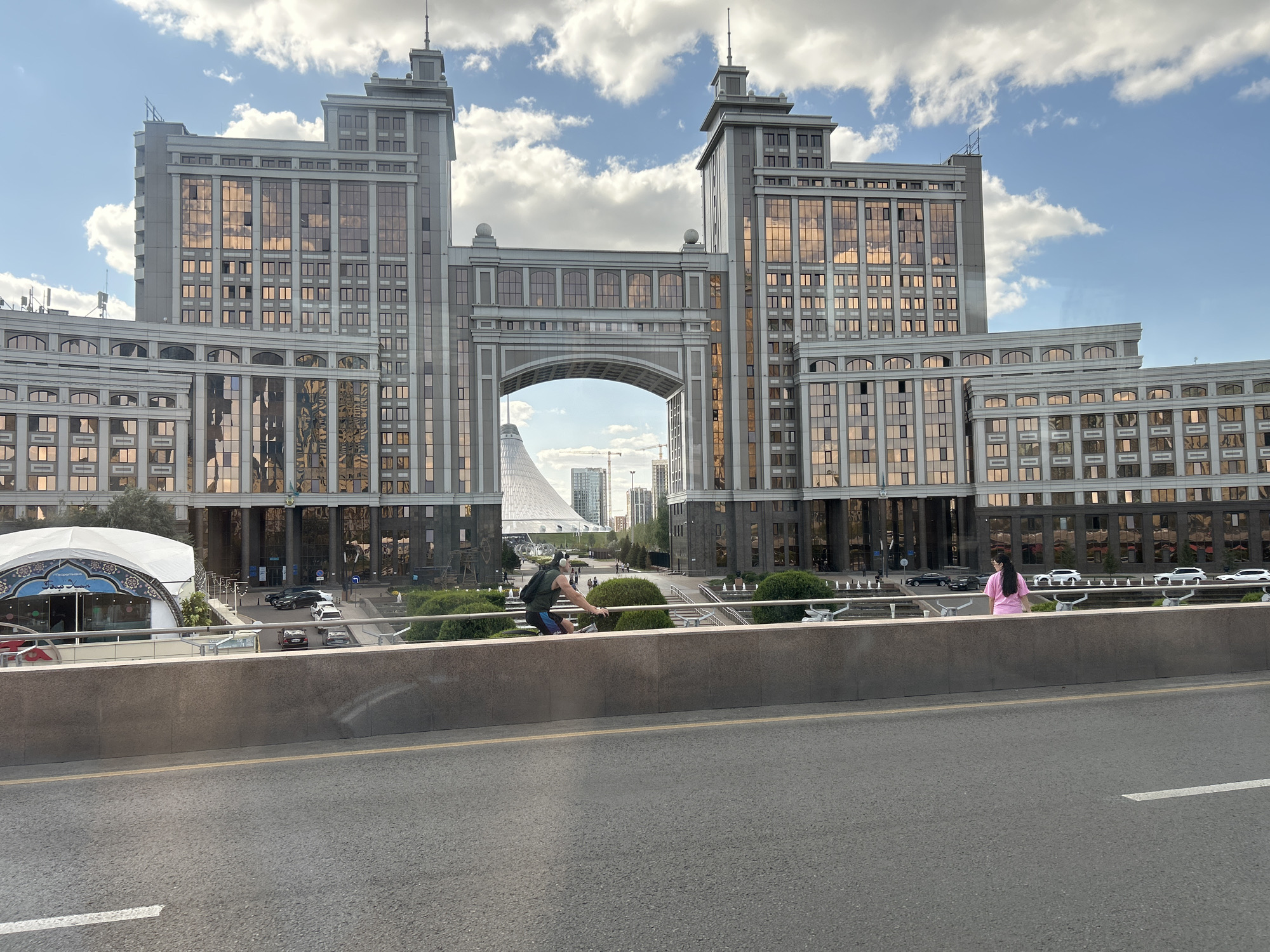 In Astana, Kazakhstan, a wide-angle, eye-level shot captures a grand architectural complex on a partly cloudy day. The buildings, constructed with a mix of grey stone and reflective gold-toned glass, are prominently featured. The central element is a large, arched gateway connecting two imposing towers. These towers, capped with spires, flank the archway, creating a symmetrical facade. Below the arch, a distinctive white, conical structure is visible in the distance, hinting at a larger urban landscape.
In the foreground, a concrete barrier runs horizontally across the lower portion of the frame, separating a roadway from the scene beyond. On top of this barrier, a man wearing headphones and a dark green tank top with blue shorts is captured in motion, appearing to be cycling or on a scooter, leaning forward as he rides. To his right, a woman in a pink t-shirt and dark pants stands facing away from the camera, looking towards the buildings. A metal railing runs behind them, along the edge of the elevated area. Beyond the barrier and railing, a manicured green space with bushes and fountains is visible, leading up to the base of the buildings. Cars are parked and driving in front of the buildings. The overall impression is one of modern urban development and public space.