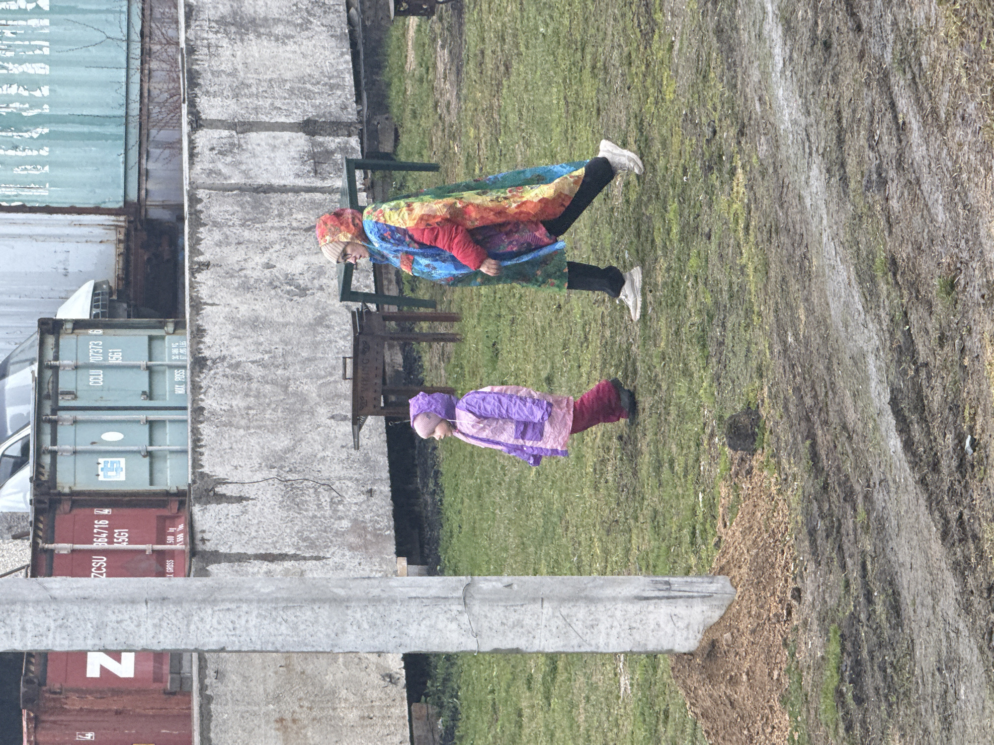 This image, likely taken in Yekaterinburg, Russia, depicts an outdoor scene featuring two people and several industrial elements. In the foreground, an adult, possibly wearing a brightly colored, rainbow-patterned hooded jacket over a red top, black pants, and white sneakers, walks or stands facing the left. They also wear glasses and a light-colored beanie. Behind and to their right, a child is visible, dressed in a purple and pink hooded jacket, red pants, and dark shoes, also facing the same direction.
The ground is a mix of green grass and muddy patches, suggesting cool and possibly wet weather. The background is dominated by large shipping containers of various colors, including light blue and red. Concrete walls, barriers, and a substantial grey concrete pole further define the industrial setting.
Visible text on the containers includes: "CCLU 07373 6", "1561", and "MAX GROSS" on a light blue container. On a red container below it, "ZCSU 364716 4", "45G1", "X. GROSS 500 kg", and "1.650" are legible. A partial "N" is seen on another red container, and "VIKING" is faintly visible elsewhere. The scene appears to capture a casual daytime moment within an urban industrial landscape.