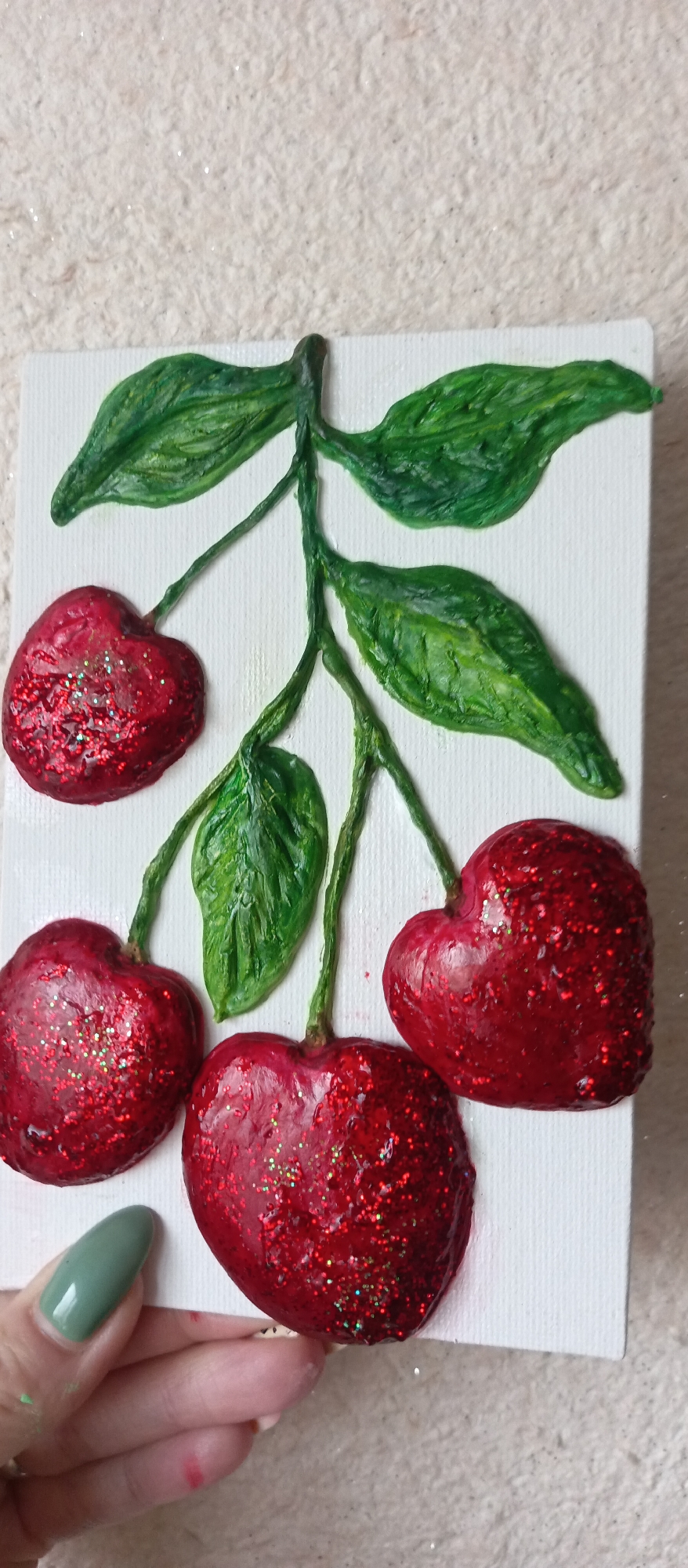 A close-up shot shows a hand holding a small, square canvas featuring an art piece depicting cherries. The canvas is white, and the cherries, leaves, and stems are three-dimensional, possibly made from clay or a similar sculpting material. The cherries are a deep red color with glitter embellishments, while the leaves and stems are green. The person's hand is holding the canvas at the bottom, and one fingernail is painted a muted green. The background is a light, textured surface.