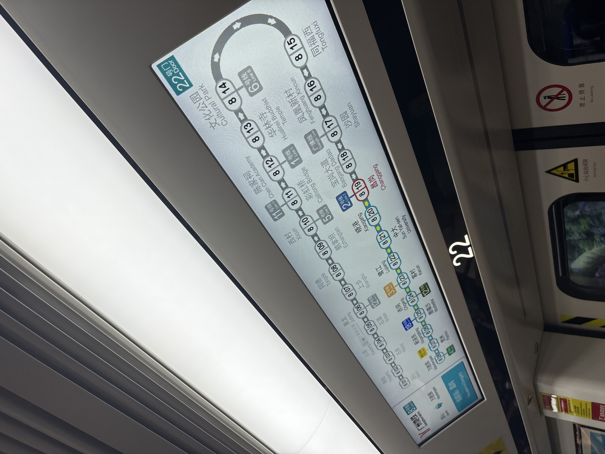 This is a medium shot, interior photograph taken from a subway train in Guangzhou, China, likely in the Baiyun District. The primary subject is a digital display board showing a subway map. The map is illuminated and displays various subway lines with station names in both Chinese and English. Several station names are visible, including "Cultural Park," "Hualinsi Temple," "Fenghuang Xincun," and "Tongfuxi" on Line 8, as well as "Chen Clan Academy" on Line 1. The train car's interior is visible in the foreground, with gray panels and overhead lighting. A large number "22" is displayed on a digital indicator above the map, possibly indicating the train number or a station identifier. There are safety signs visible on the train doors, including a "Warning Gap" sign with a yellow triangle and a "No Leaning" sign with a red circle and a diagonal line over a person. The environment is that of a modern public transportation vehicle. No people are clearly discernible within the frame. The angle of the photograph suggests it was taken from a seated or standing position within the train, looking upwards at the display.
