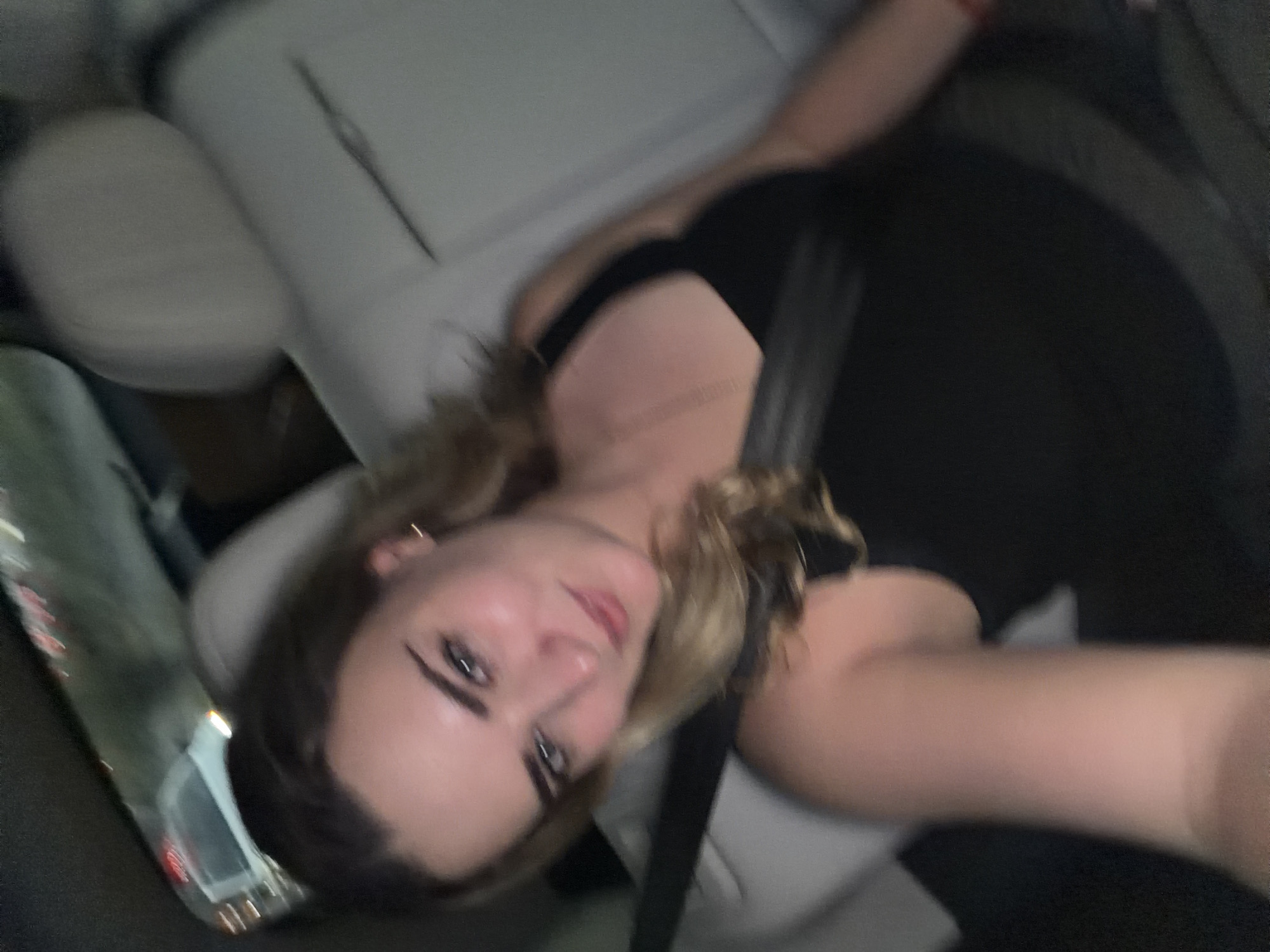 The image features a close-up portrait of a young woman inside what appears to be a car, likely captured as a selfie or a spontaneous shot. She is seated on a light grey or beige car seat, with a black seatbelt visible diagonally across her chest. Her head is tilted, and she looks directly into the camera with a subtle, calm expression and a hint of a smile. She has medium-length, wavy hair that is blonde or light brown in color. Her facial features are accented by defined eyebrows, dark eyes with winged eyeliner, and naturally toned or lightly colored lips. She is wearing a dark, possibly black, top, a delicate gold-colored chain necklace, and small hoop earrings. The background behind her and visible through the vehicle's window is softly blurred, indicative of either motion or low-light conditions. Reflections of external lights, such as streetlights or other car lights, are noticeable in the window area, suggesting the photograph was taken during the evening or at night. The scene captures a moment within a vehicle, possibly while traveling through San Bernardino Tlaxcalancingo, Mexico. No discernible text is visible in the image.
