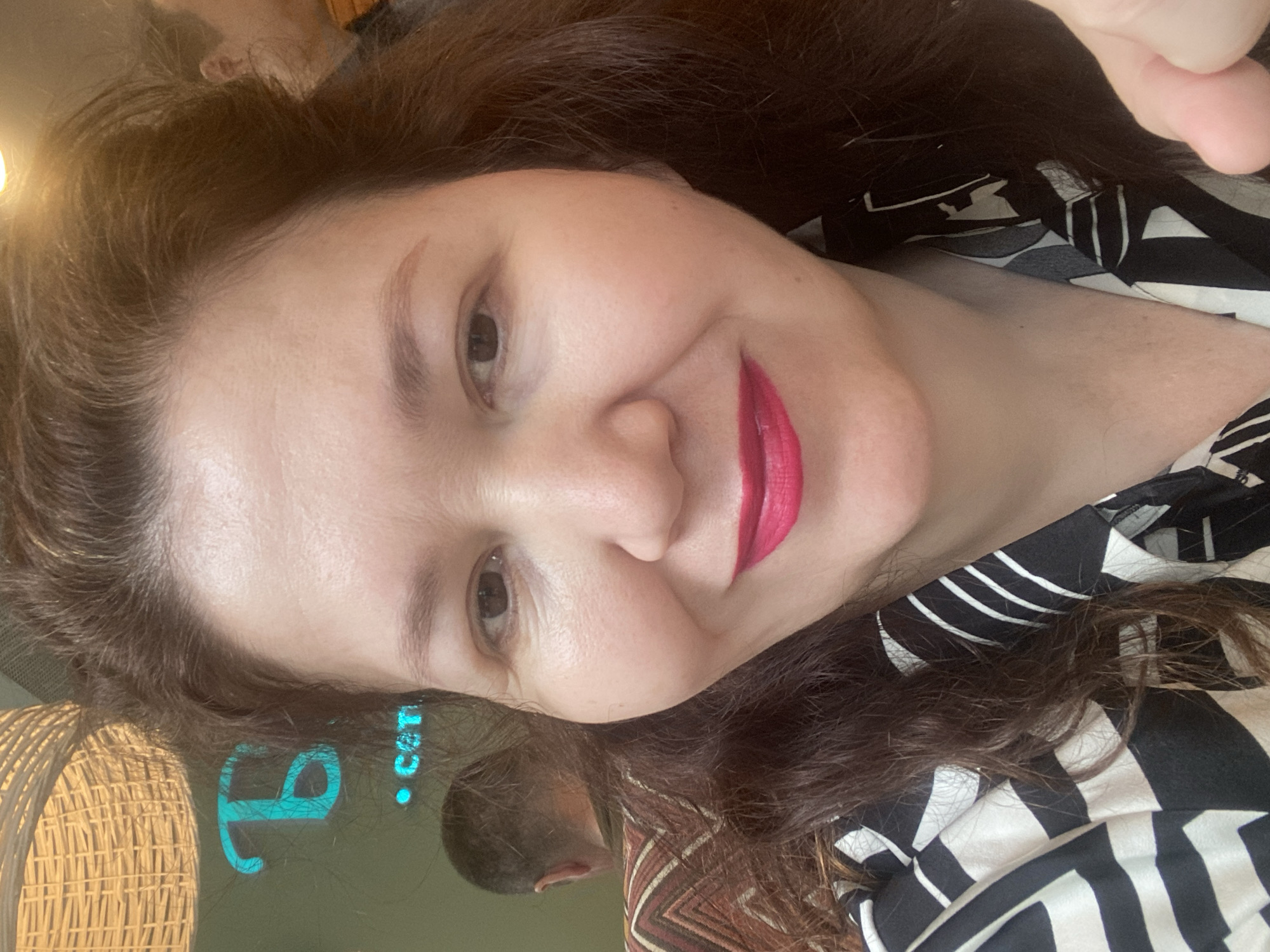 A close-up, selfie-style, eye-level shot captures a woman from the chest up, looking directly at the camera with a slight smile. Her face is illuminated, highlighting her fair skin, brown eyes, and bright pink lipstick. Her dark brown hair is slightly tousled and frames her face. She is wearing a black and white patterned shirt. In the background, a woven light fixture hangs from above, and on the left, a neon turquoise sign with Cyrillic lettering is visible, along with the word "CBT" in smaller text. To her left, the back of a man's head with short hair is partially visible, and further in the background, another person is visible. The setting appears to be an indoor establishment, possibly a cafe or restaurant. The neon sign suggests the location may be in Ufa, Russia, as "CBT" could be part of a business name or abbreviation common in the city.
