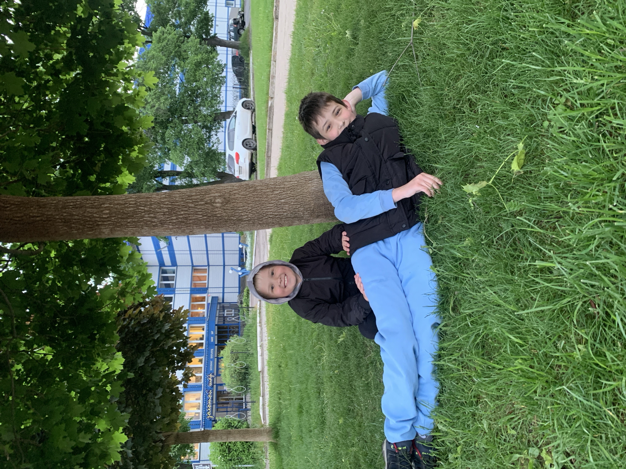 Two boys are lying on a grass field, back-to-back, near a tree. The boy on the right is wearing a light blue long-sleeved shirt, a black vest, and light blue pants. The boy on the left is wearing a black hooded jacket and light blue pants. The background shows a building and cars parked on the street. The building features blue and white striped walls with lighted windows. The location is identified as Chaikovsky, Russia.